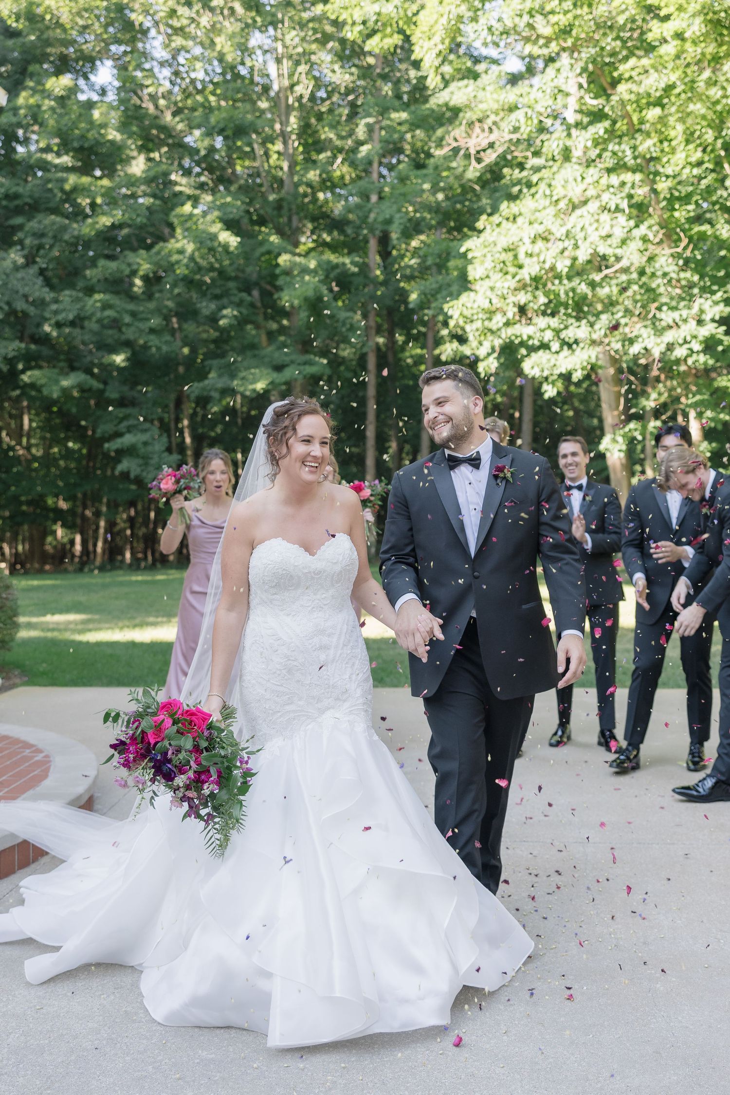 Wedding party throws pink flower petals at newlyweds during outdoor garden celebration.