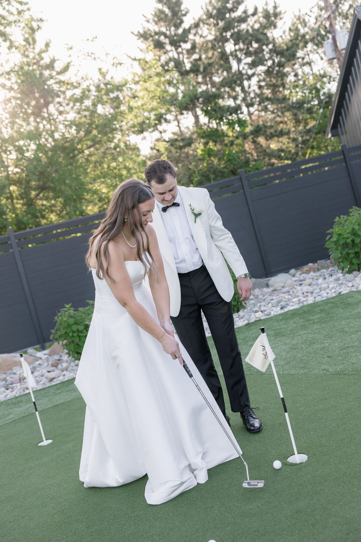 Bride and groom playing mini golf on putting green, groom in white tuxedo guiding bride in wedding gown.