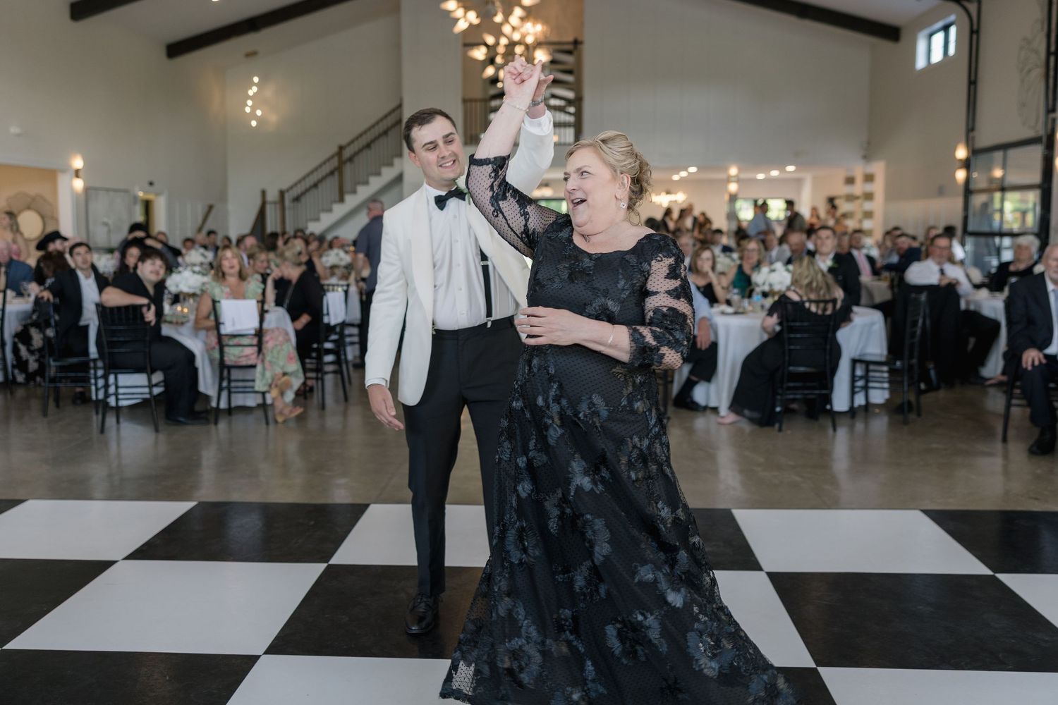A couple elegantly dances on a black and white checkered floor at a wedding reception in a bright venue.