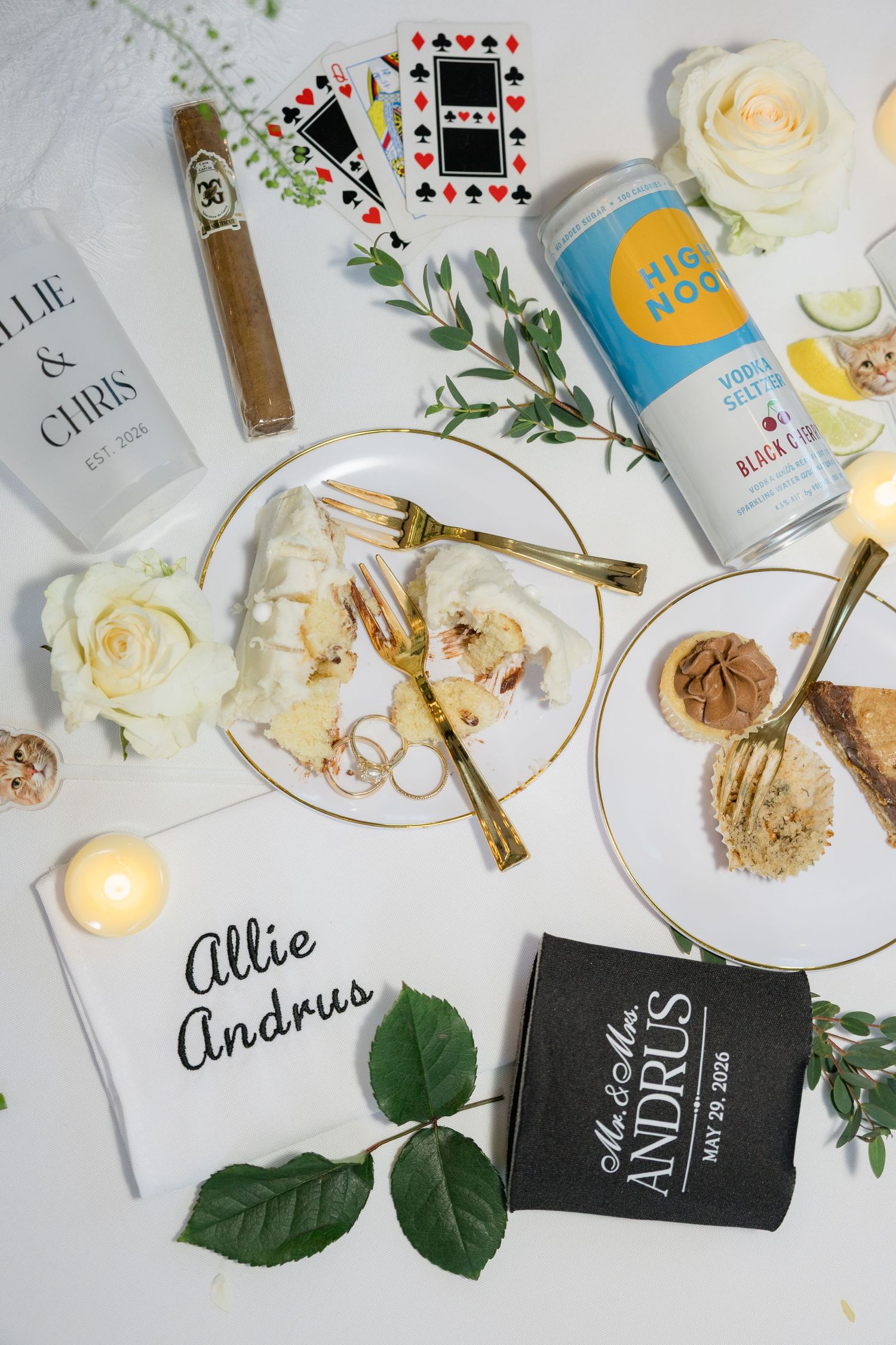 Wedding flat lay with personalized napkins, koozies, playing cards, cigar, High Noon drink, cake slices, and white roses.