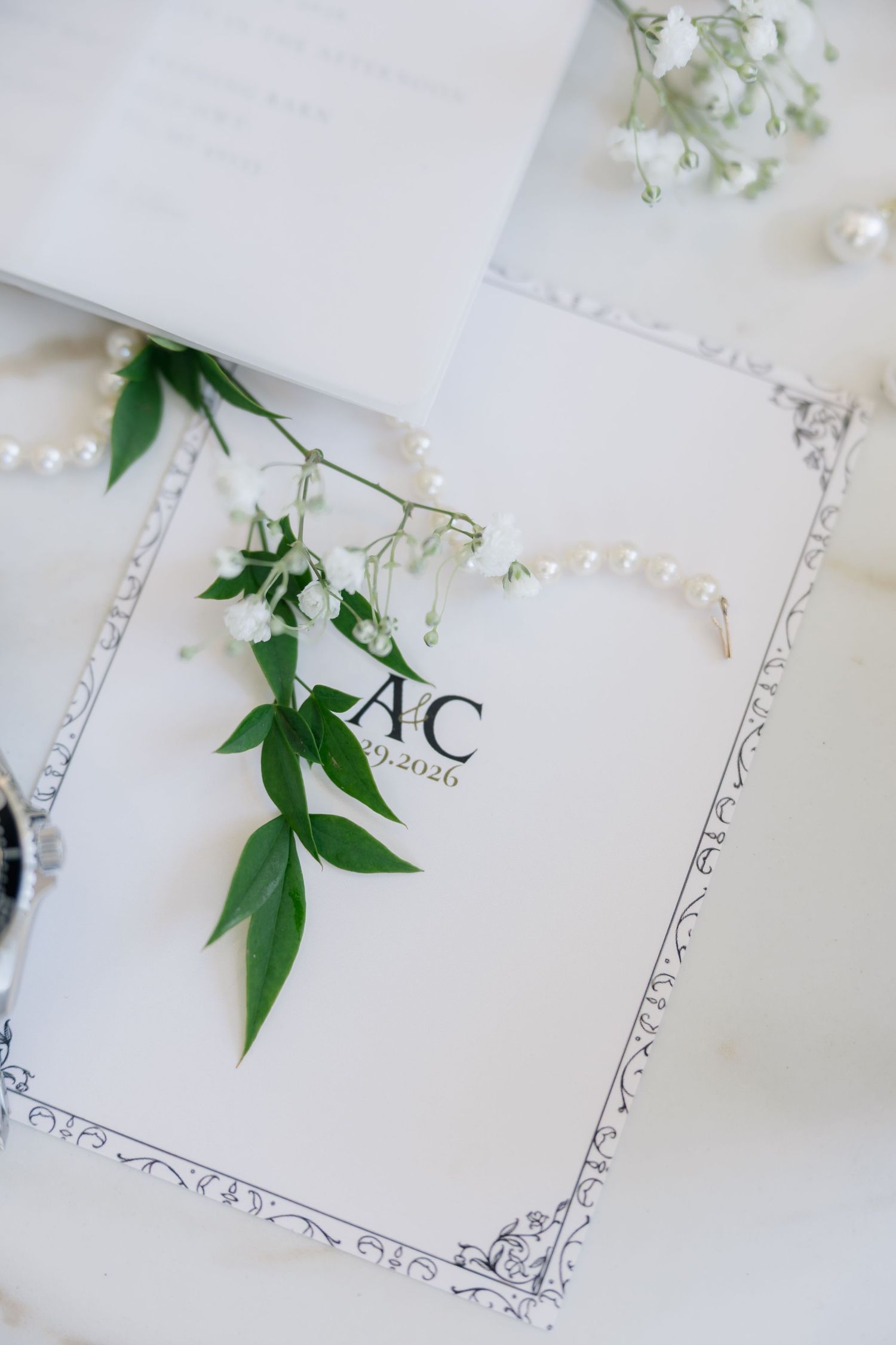 Elegant wedding stationery with monogram AC 2026, pearl necklace, and green floral sprigs on white marble.