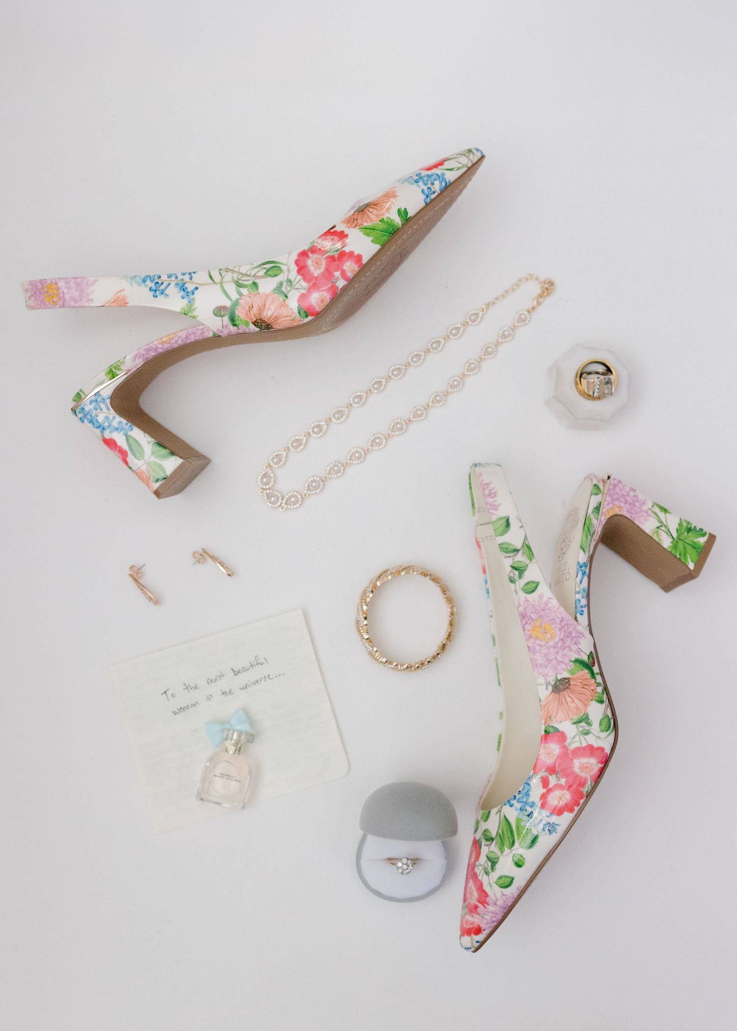 Overhead shot of floral wedding shoes with jewelry and accessories on white background.