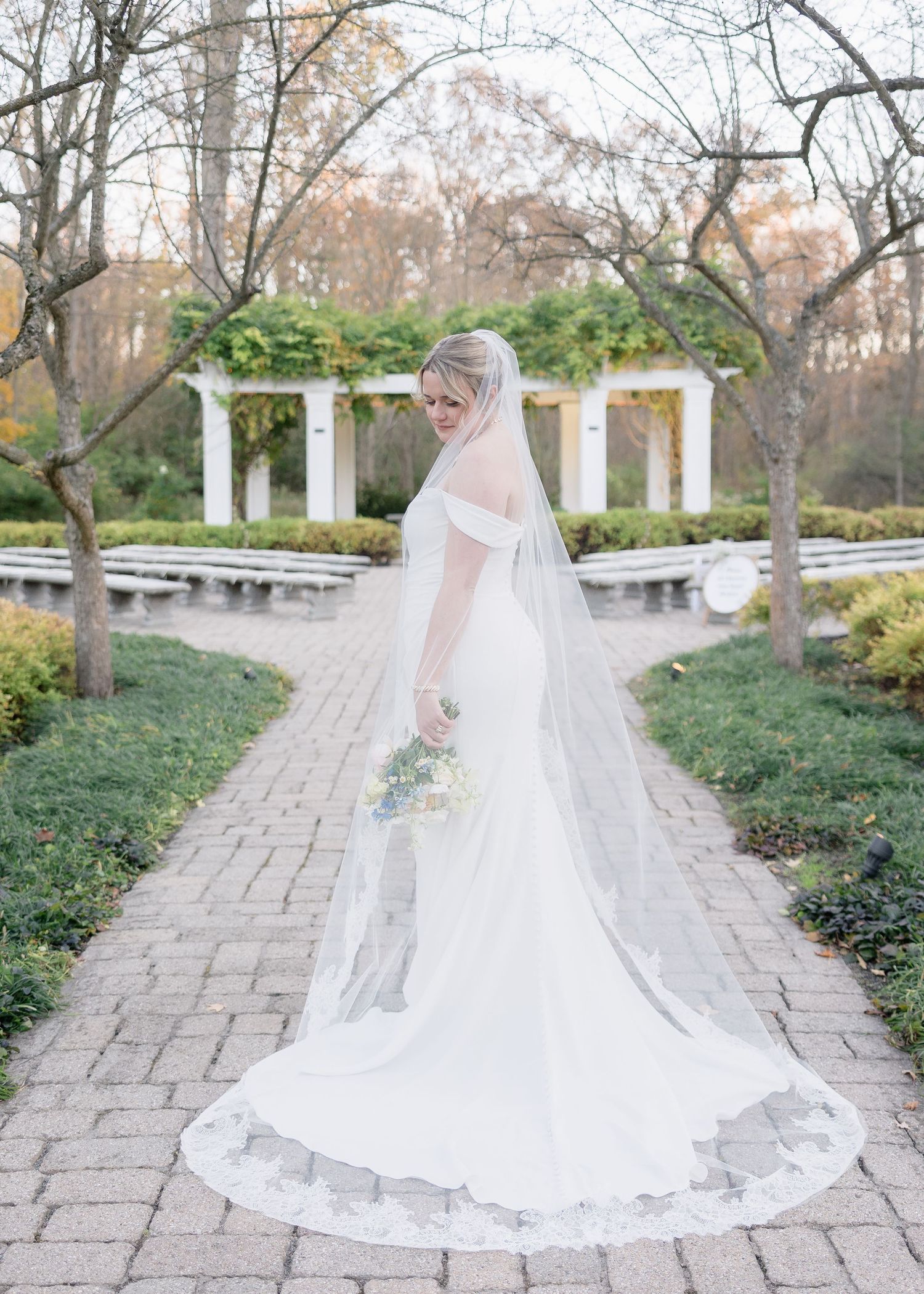 A bride in a flowing white dress and veil poses on a stone path in a garden with white columns.