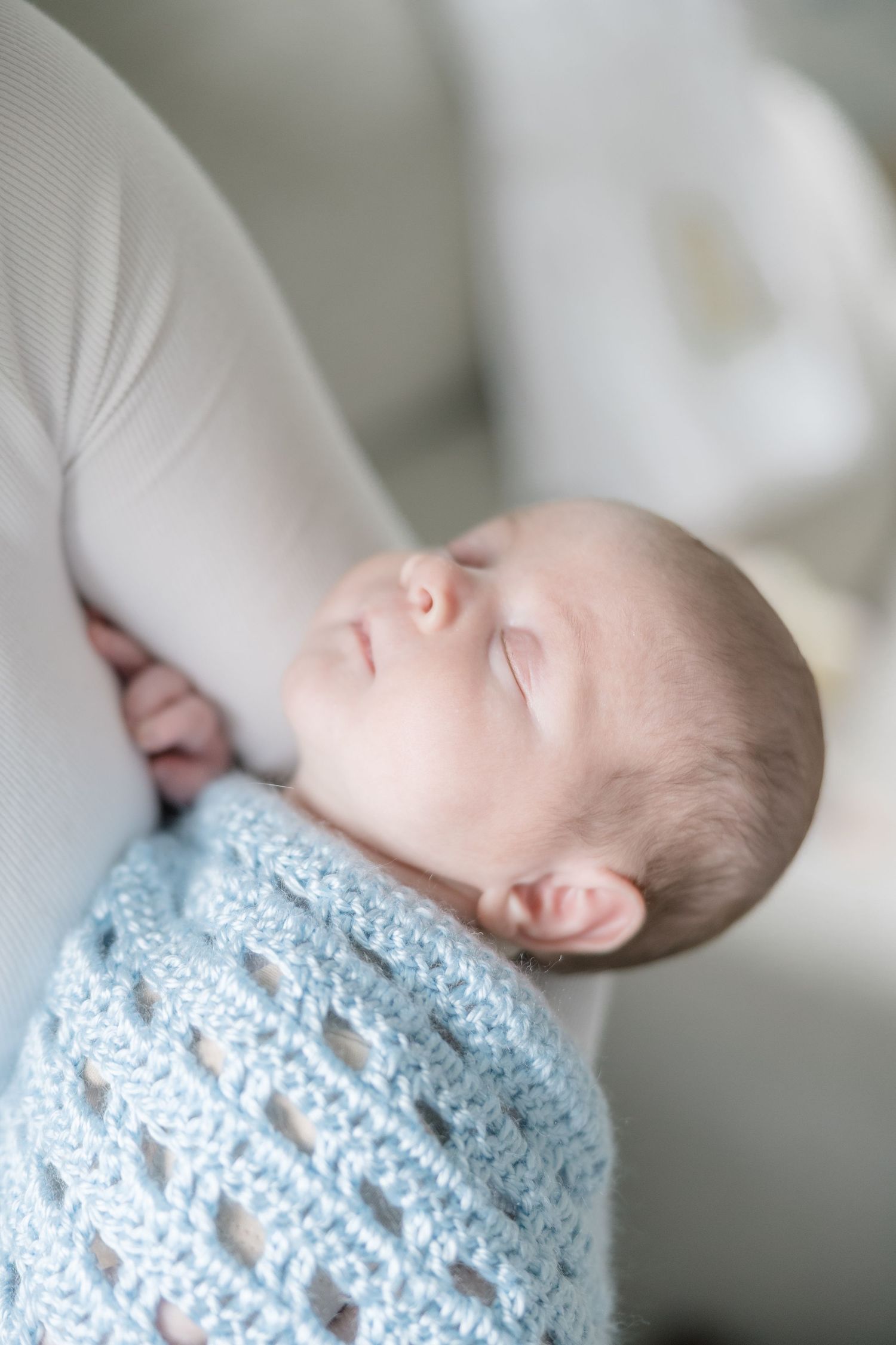 A sleeping infant wrapped in a soft blue knitted blanket rests peacefully against white fabric.