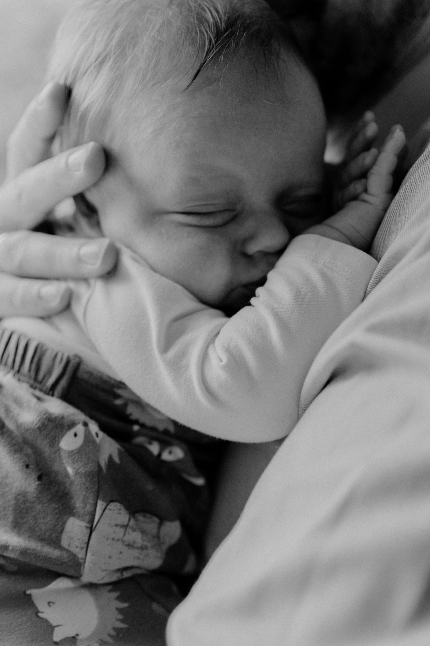 A sleeping infant rests peacefully in black and white, nestled against soft bedding with a gentle hand touching their face.