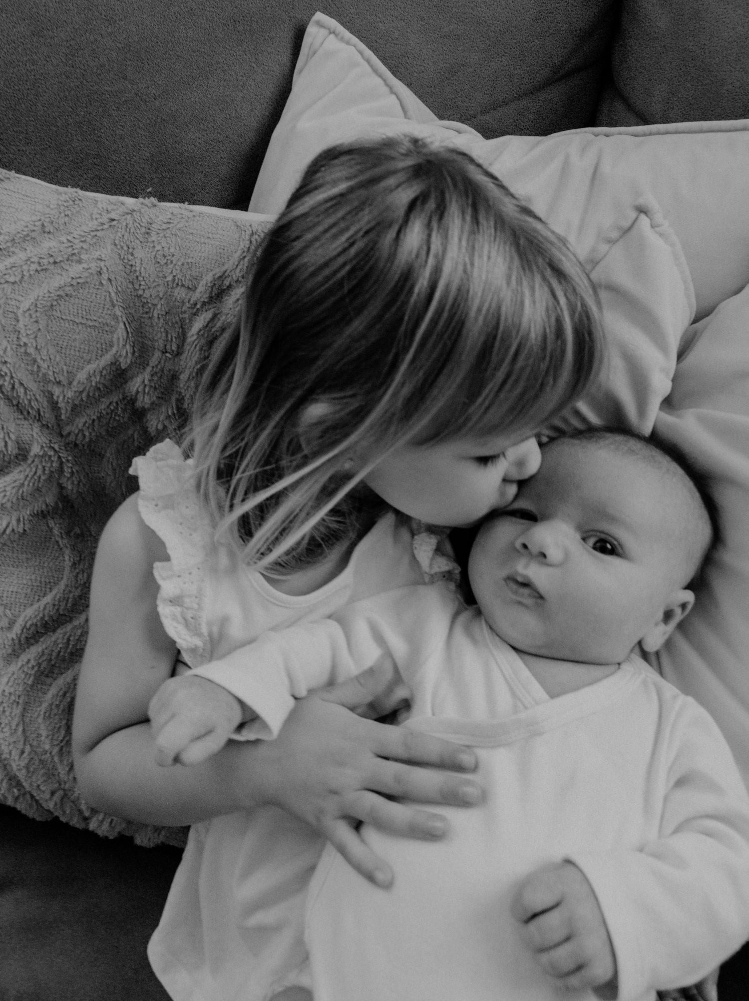 Sweet black and white photograph of two siblings bonding on a couch as older sister gently kisses baby's cheek.
