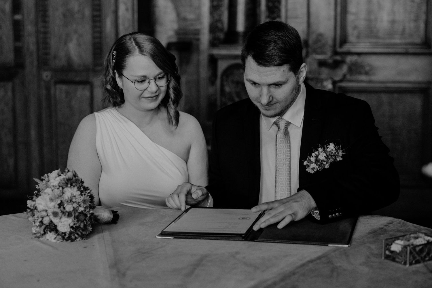 A couple signs their marriage documents at a formal ceremony with a bouquet on the table.