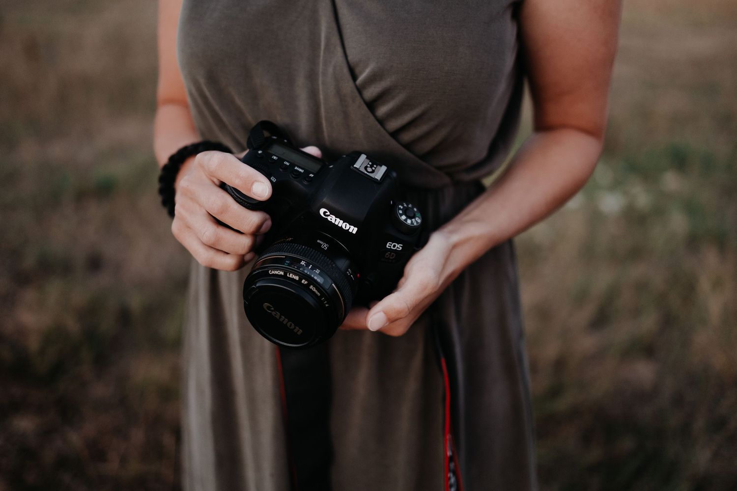 A person wearing a brown dress holds a Canon DSLR camera in their hands against a blurry outdoor background.