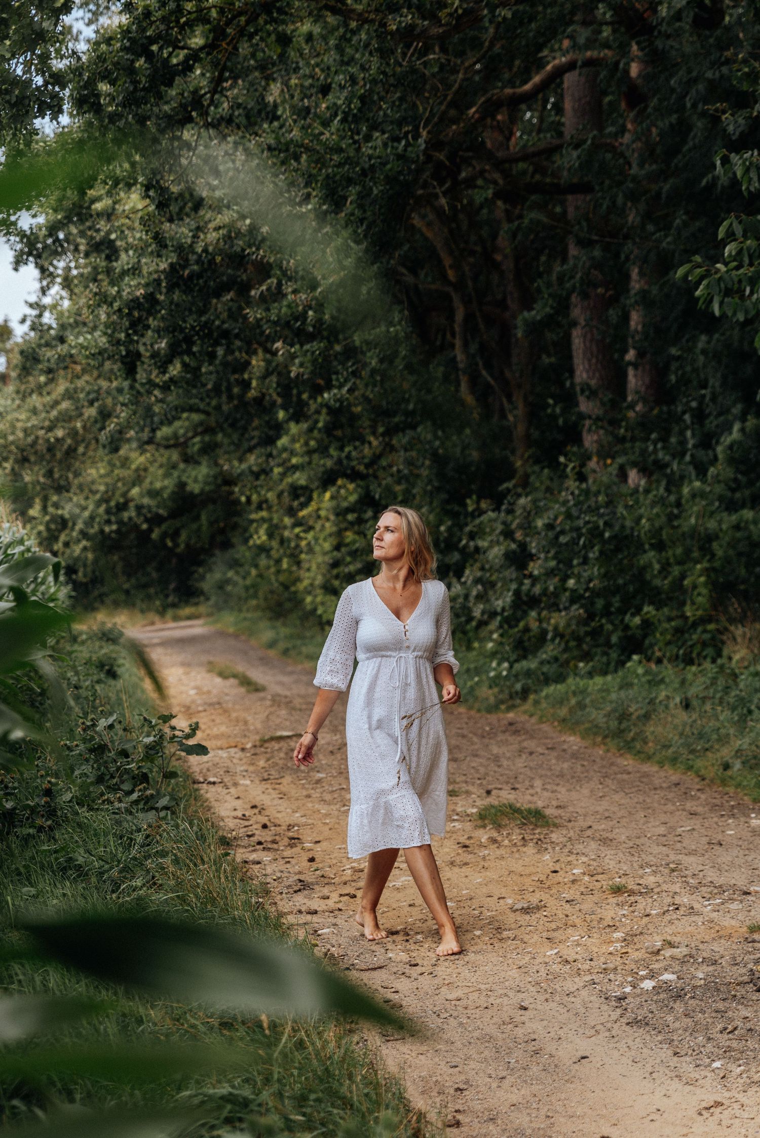 Someone in a white summer dress walks barefoot along a winding dirt path through lush green forest scenery.