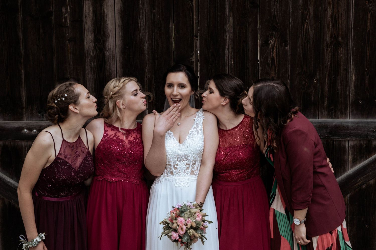 Bridesmaids in burgundy dresses surround bride in white lace gown while holding pink bouquet outdoors.