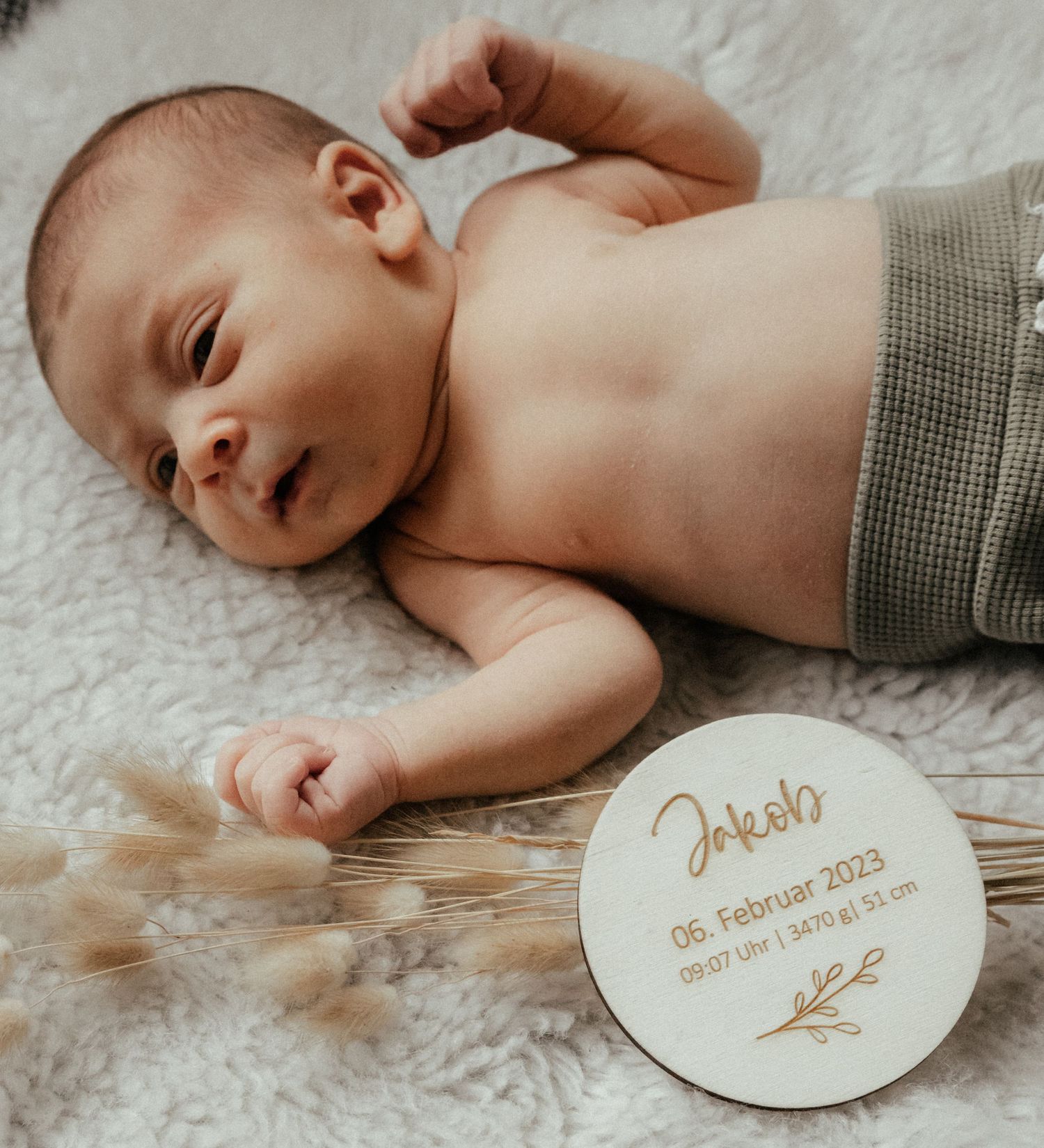 Newborn baby lying on white blanket with personalized wooden name plaque showing birth details.