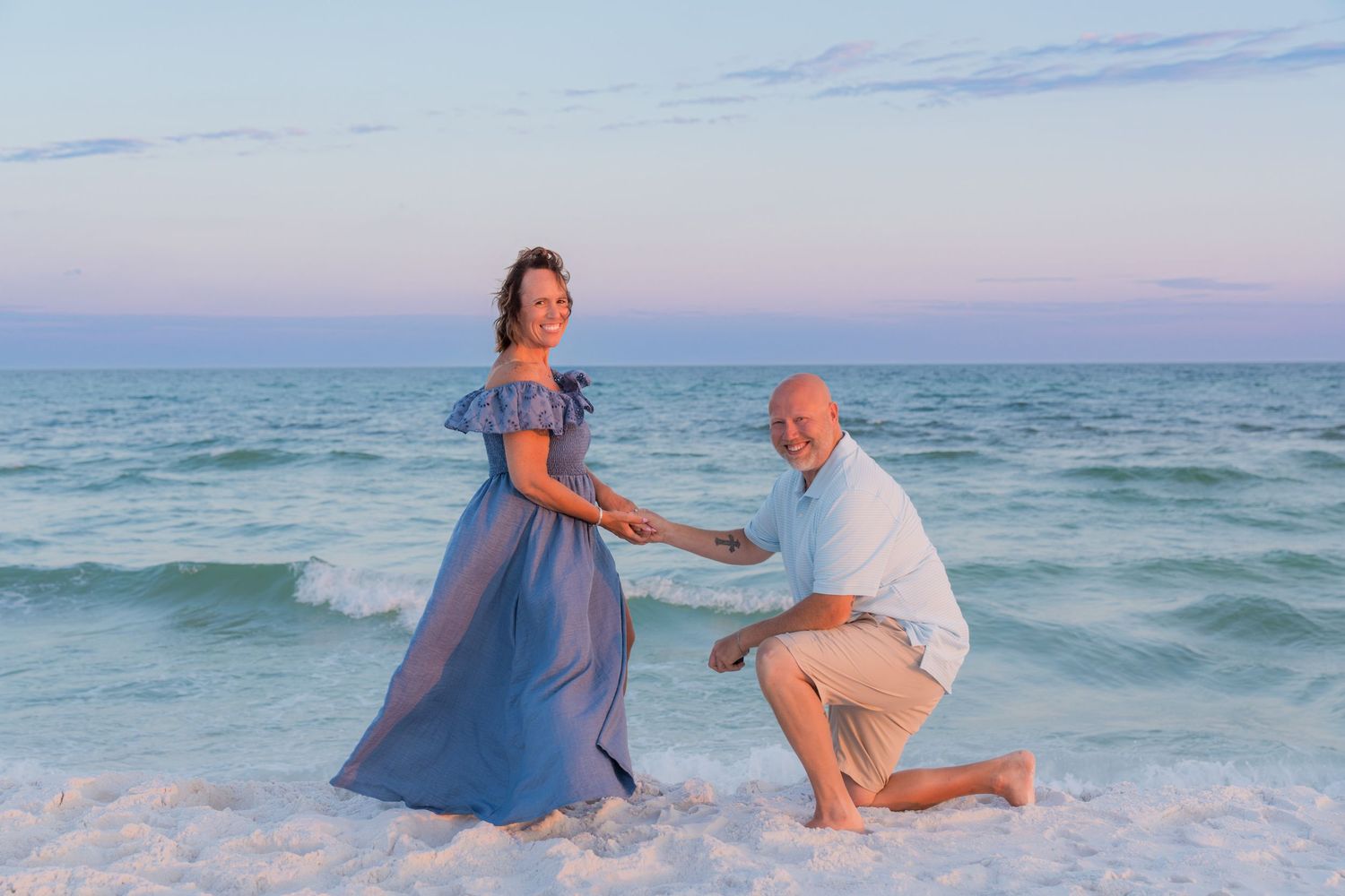 Man kneeling on beach proposing to woman in blue dress at sunset with ocean waves in background.