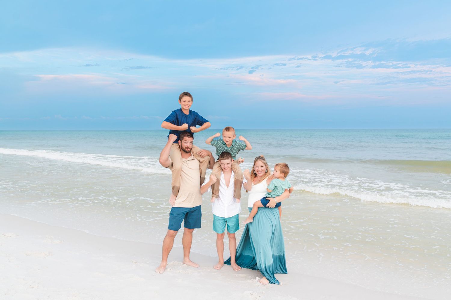 Family of six poses on a white sandy beach at sunset, with two boys on shoulders, wearing coordinated blue outfits.