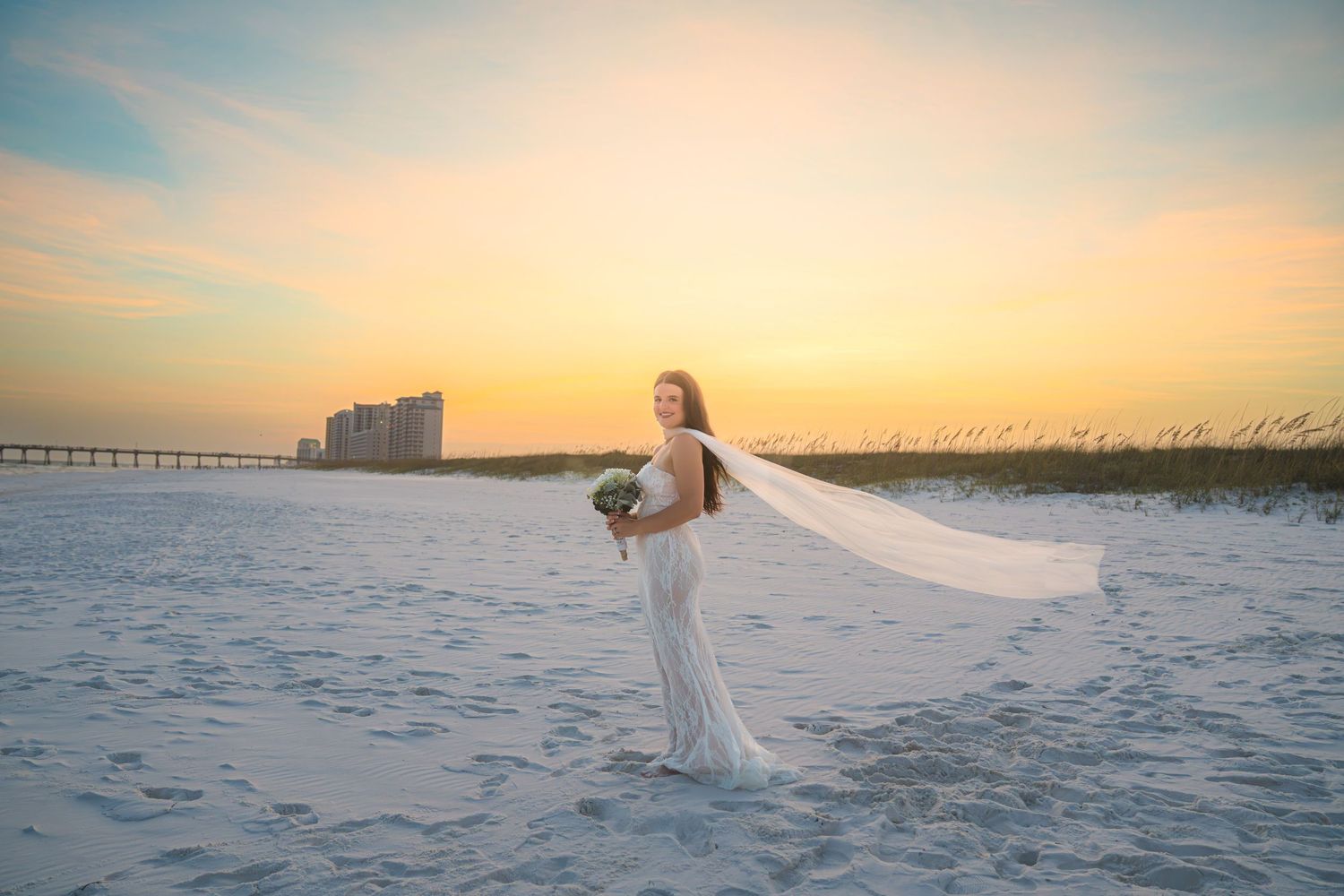 Bride in white gown and flowing veil poses on sandy beach at sunset with bouquet, pier and dunes in background.