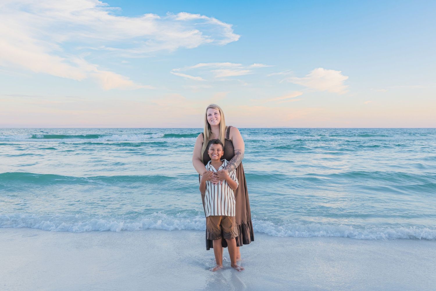 Woman and child standing together on a sandy beach with turquoise ocean waves and pastel sunset sky.