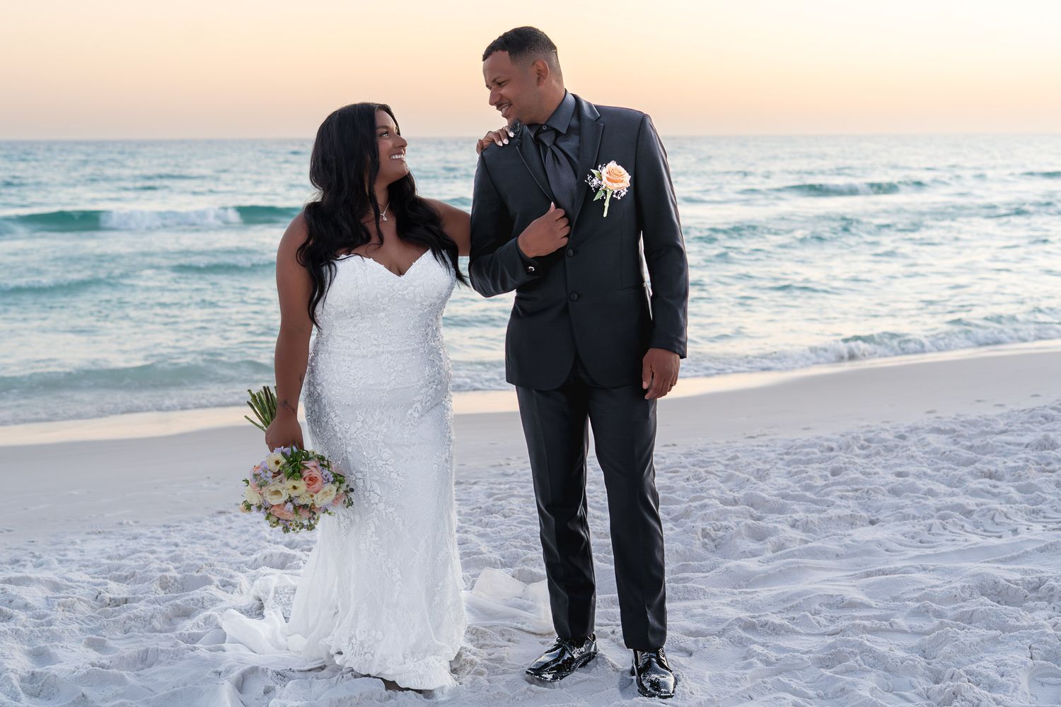 Bride and groom share a romantic moment on a sandy beach at sunset, bride holding a bouquet in a white gown.