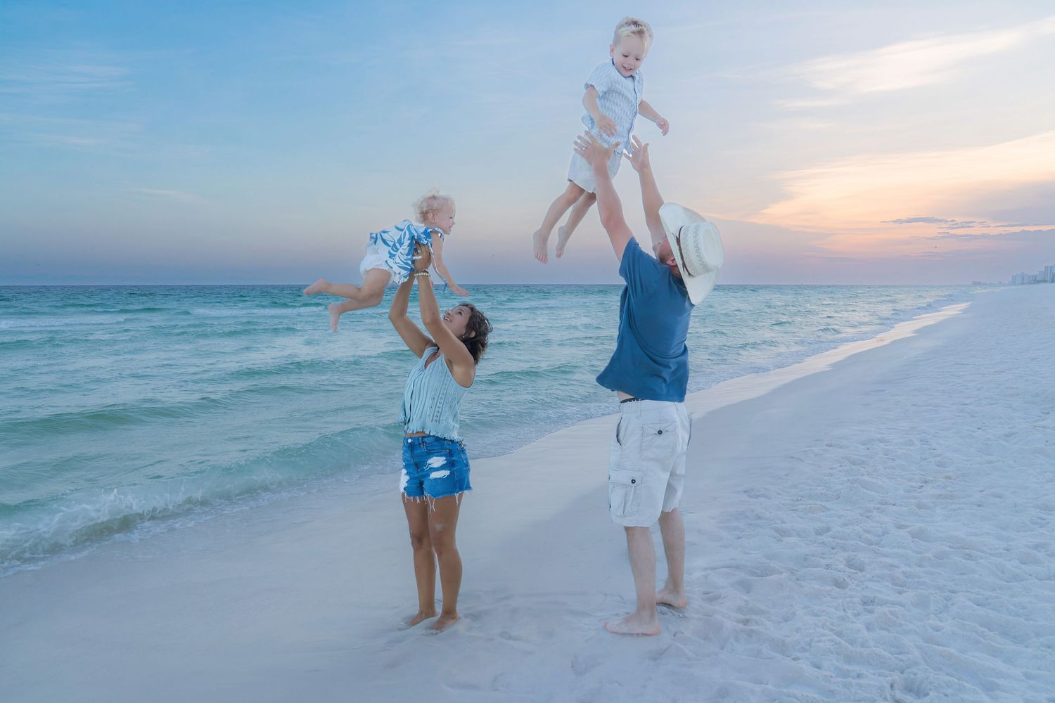 Family joyfully tossing two babies in the air on a beautiful beach at sunset with turquoise waves.