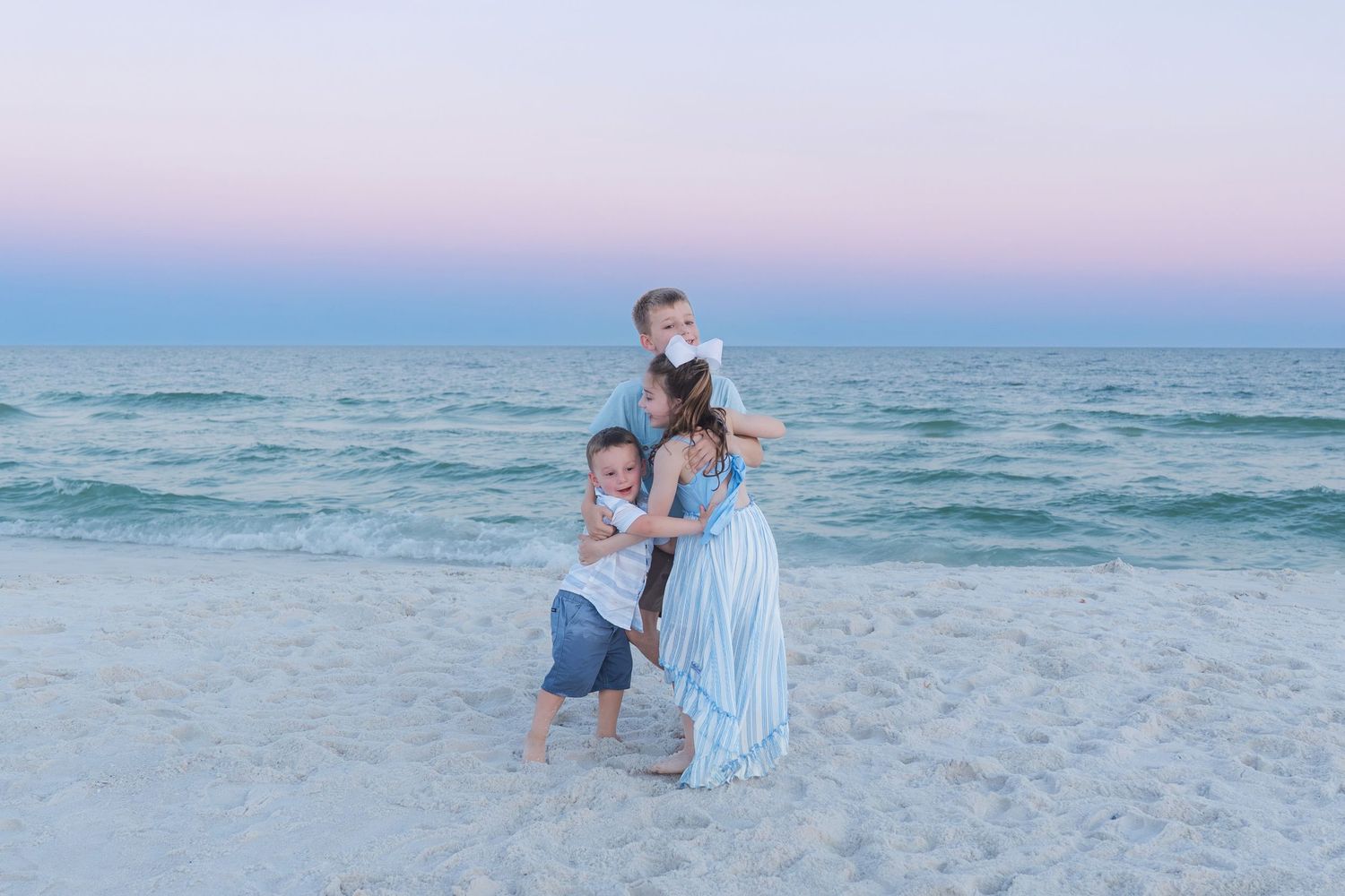 Mother in blue dress hugging two children on beach at sunset with pastel pink and blue sky backdrop.