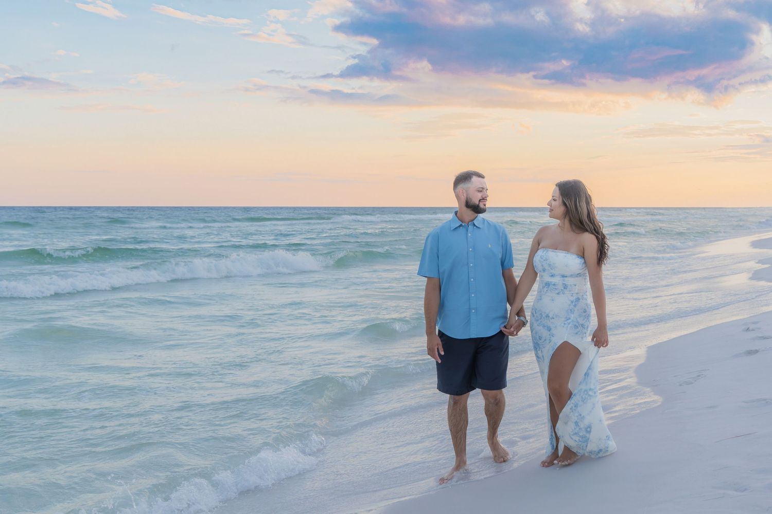 Couple holding hands walking on a sandy beach at sunset with turquoise waves and pastel sky.