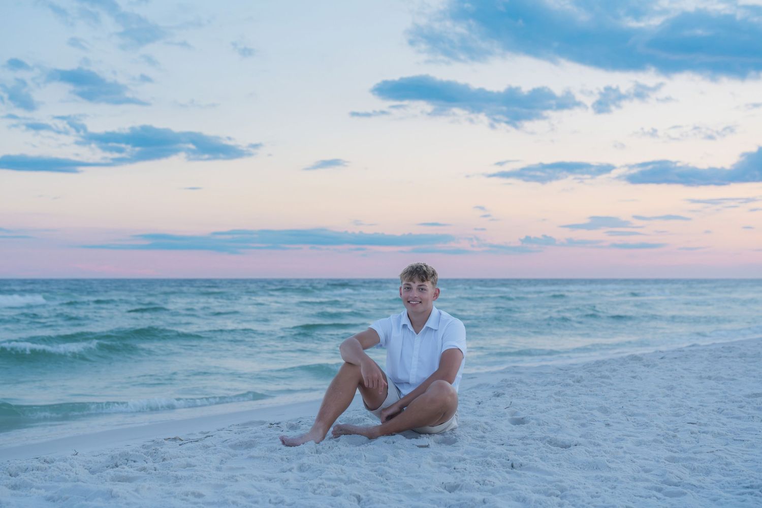 Young man sitting cross-legged on white sand beach at sunset with pastel sky and turquoise ocean behind him.