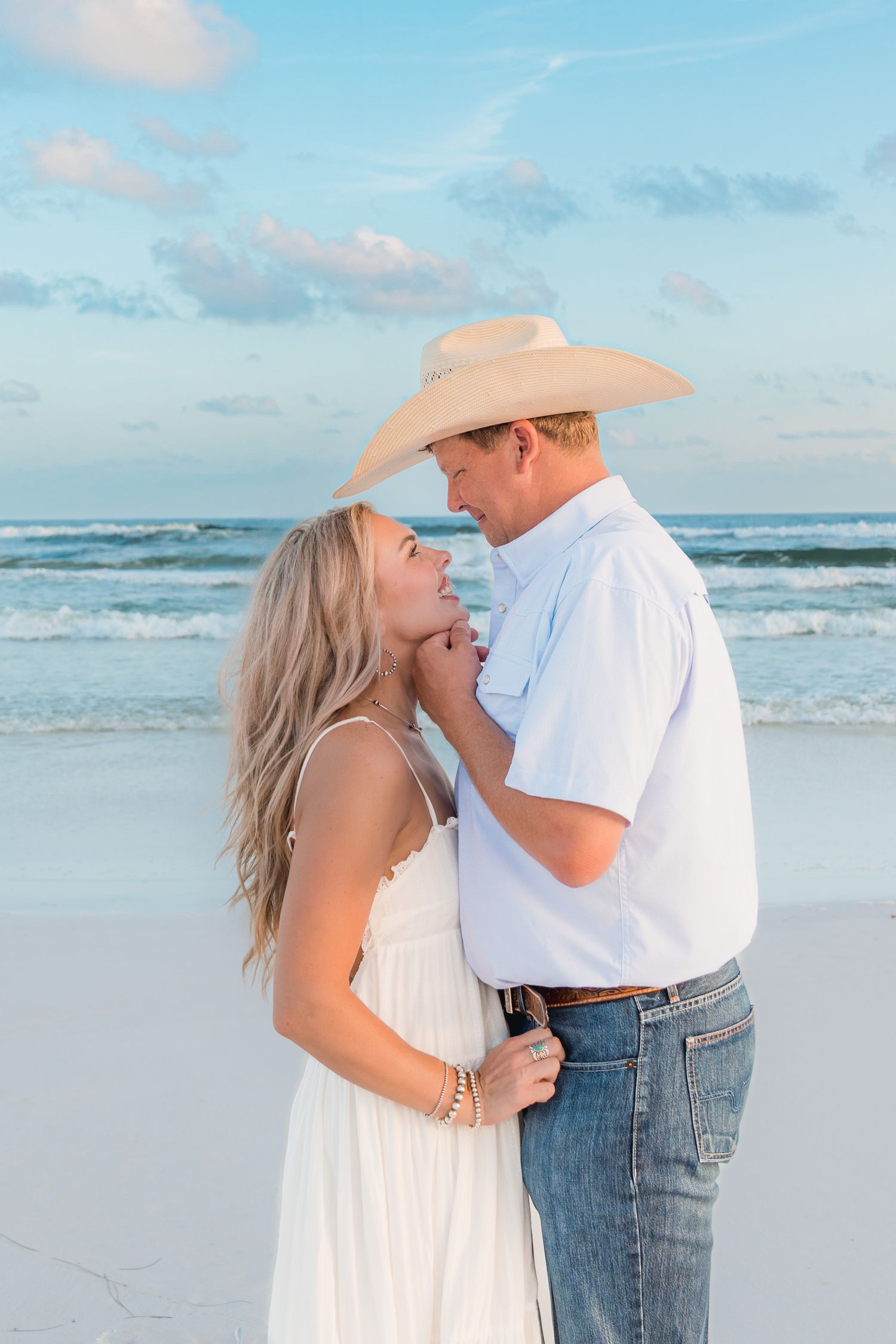 Couple in white dress and cowboy hat share a romantic moment on a beach with ocean waves behind them.