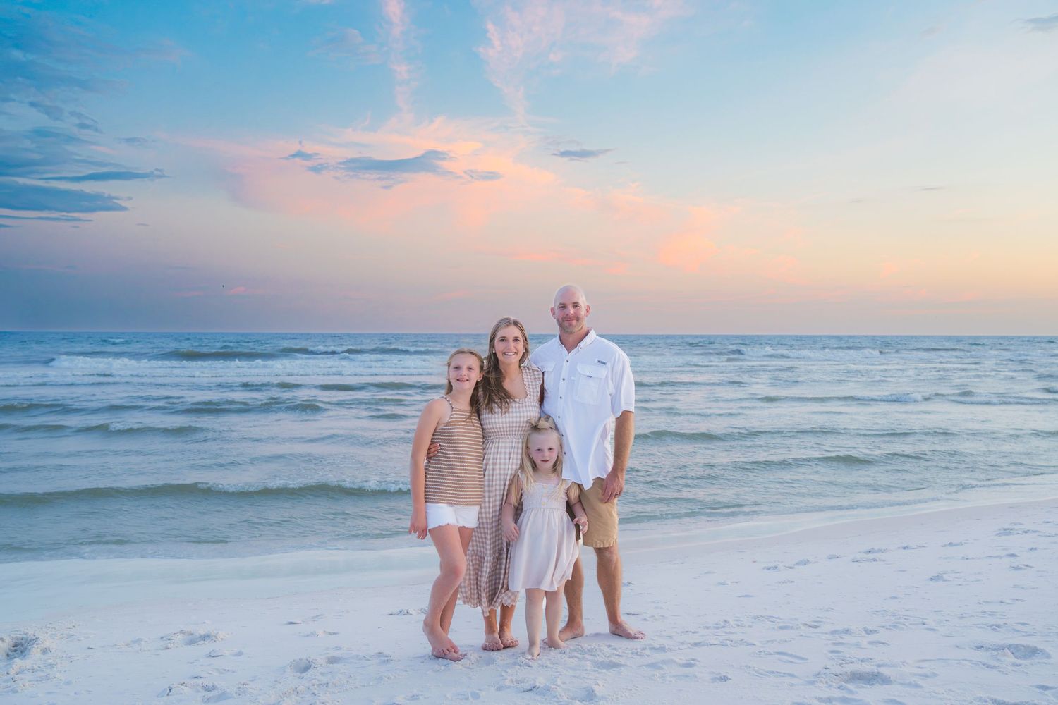 Family of four posing on a white sand beach at sunset with pastel skies and calm ocean waves.