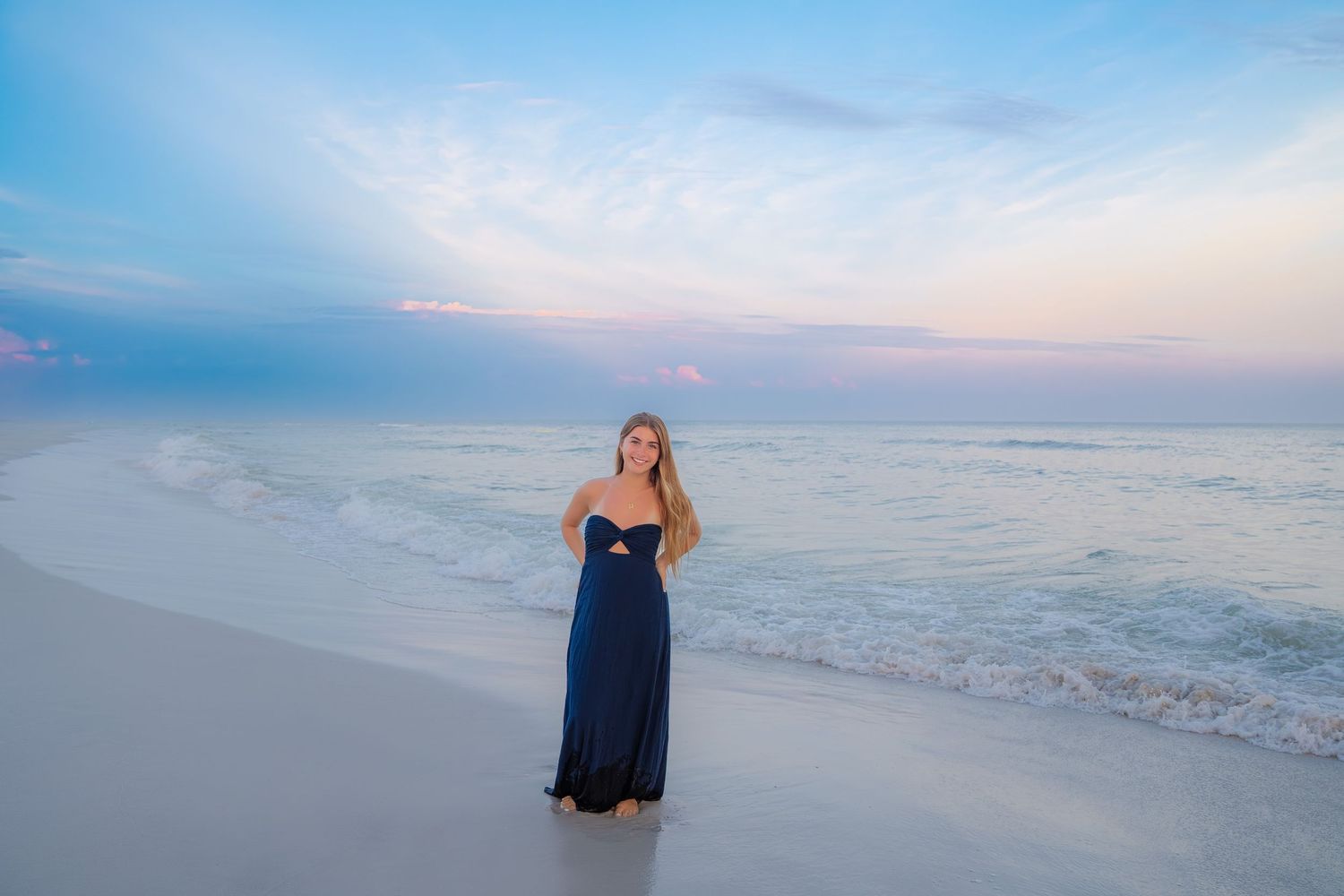Woman in navy maxi dress posing on white sand beach at sunset with pastel sky and ocean waves.