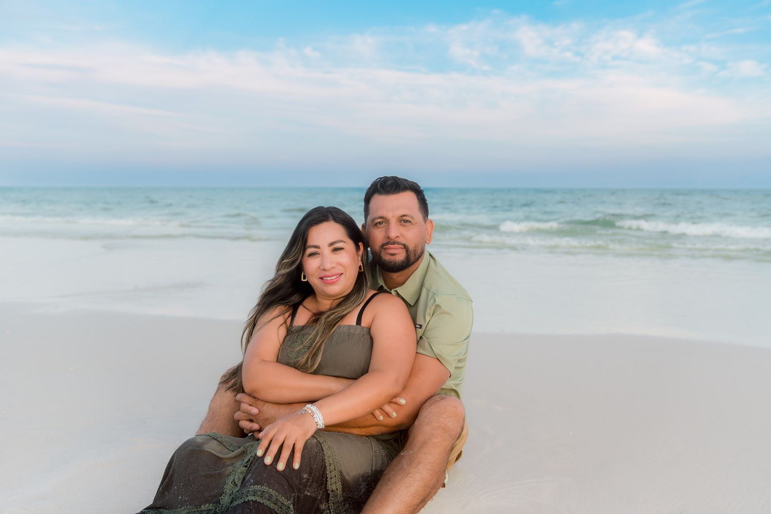 Couple sitting together on white sand beach with turquoise ocean waves and blue sky backdrop.