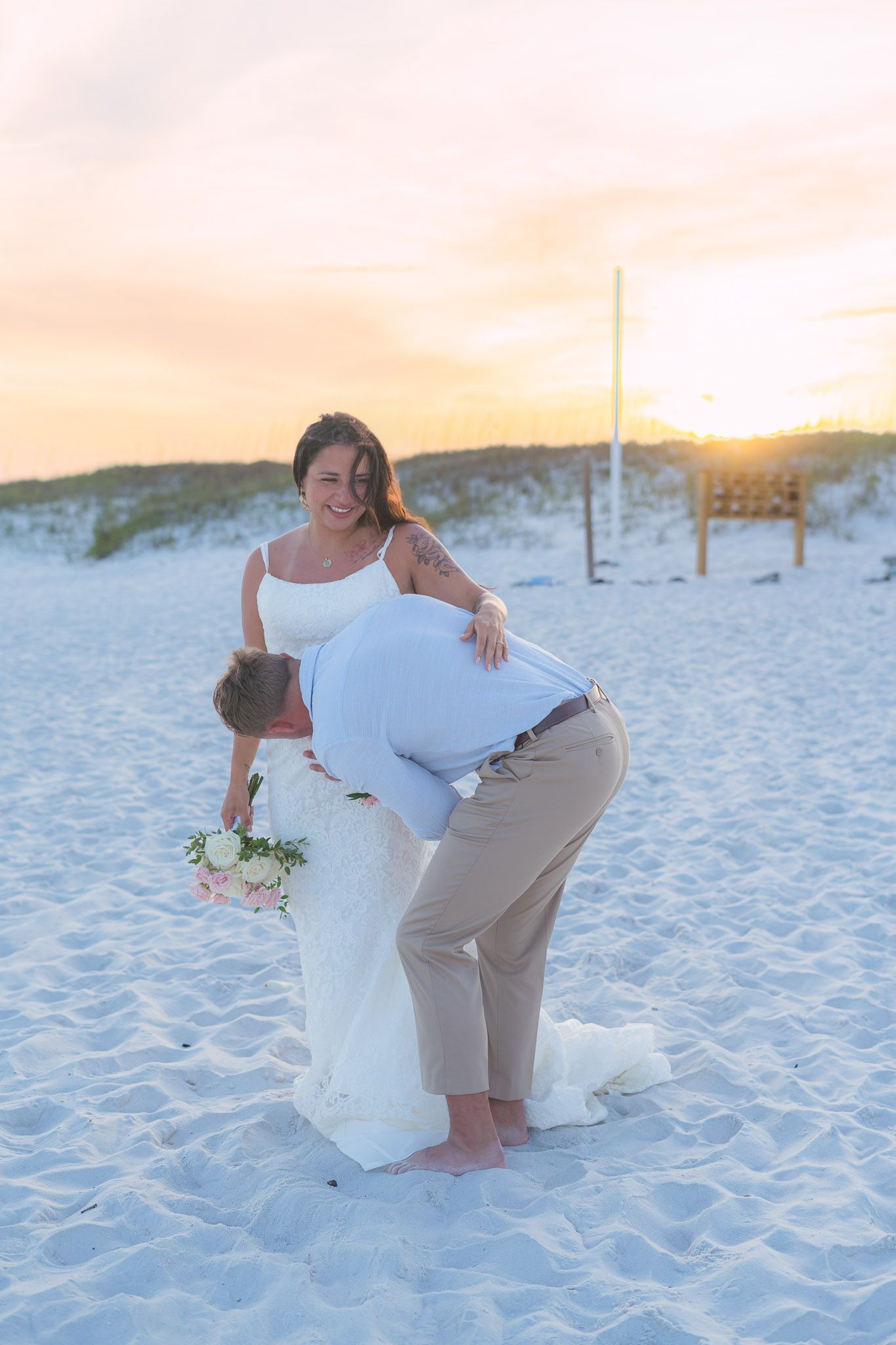 Bride and groom share a romantic dip kiss on a white sand beach at sunset during their wedding celebration.