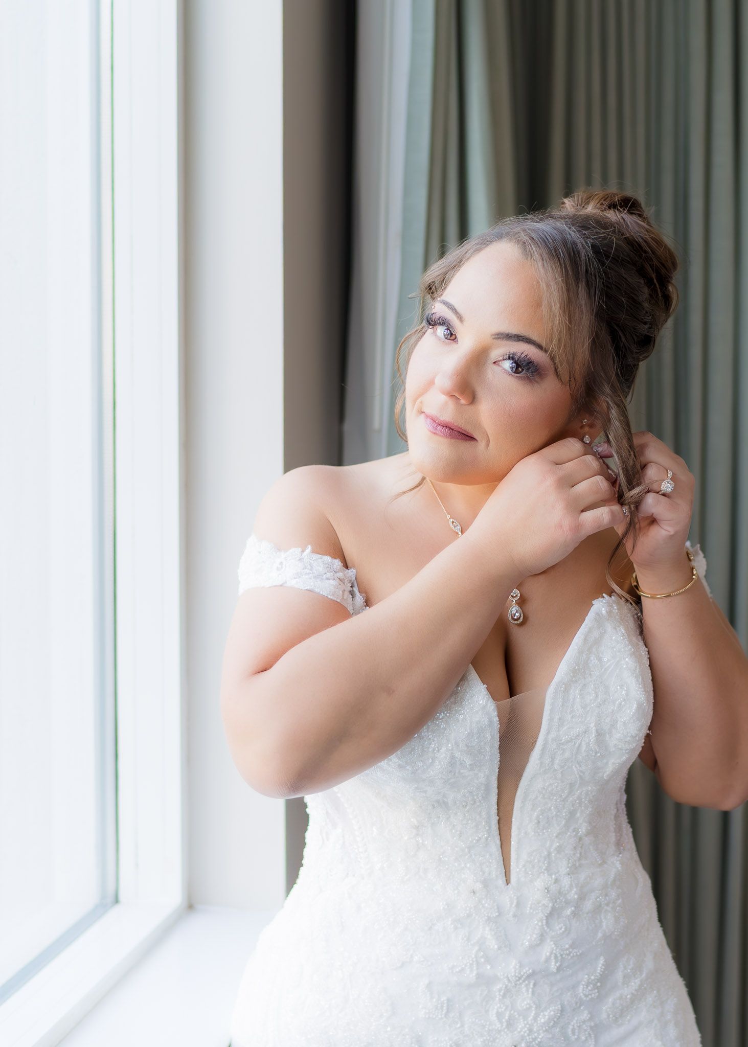 Bride in lace off-shoulder gown putting on earrings by window, wearing gold jewelry and elegant updo hairstyle.