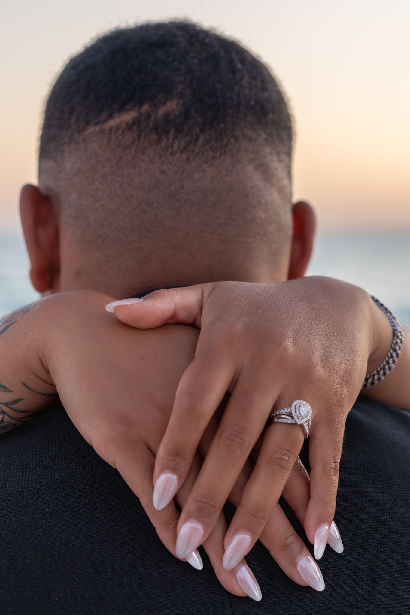 A woman displays her diamond engagement ring with white nails while embracing a man at sunset.
