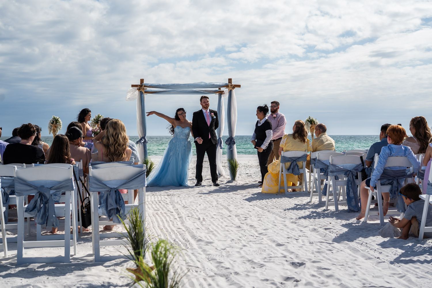 Beach wedding ceremony with couple standing at rustic wooden arch on white sand beach.