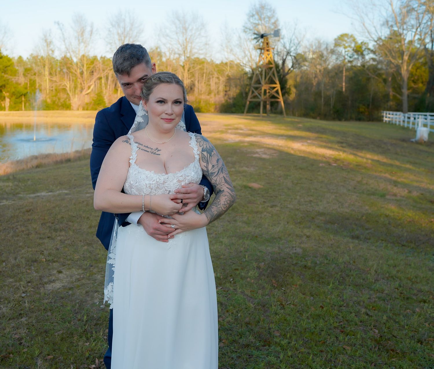 Groom embraces bride from behind in a sunlit outdoor field with a windmill and pond in background.