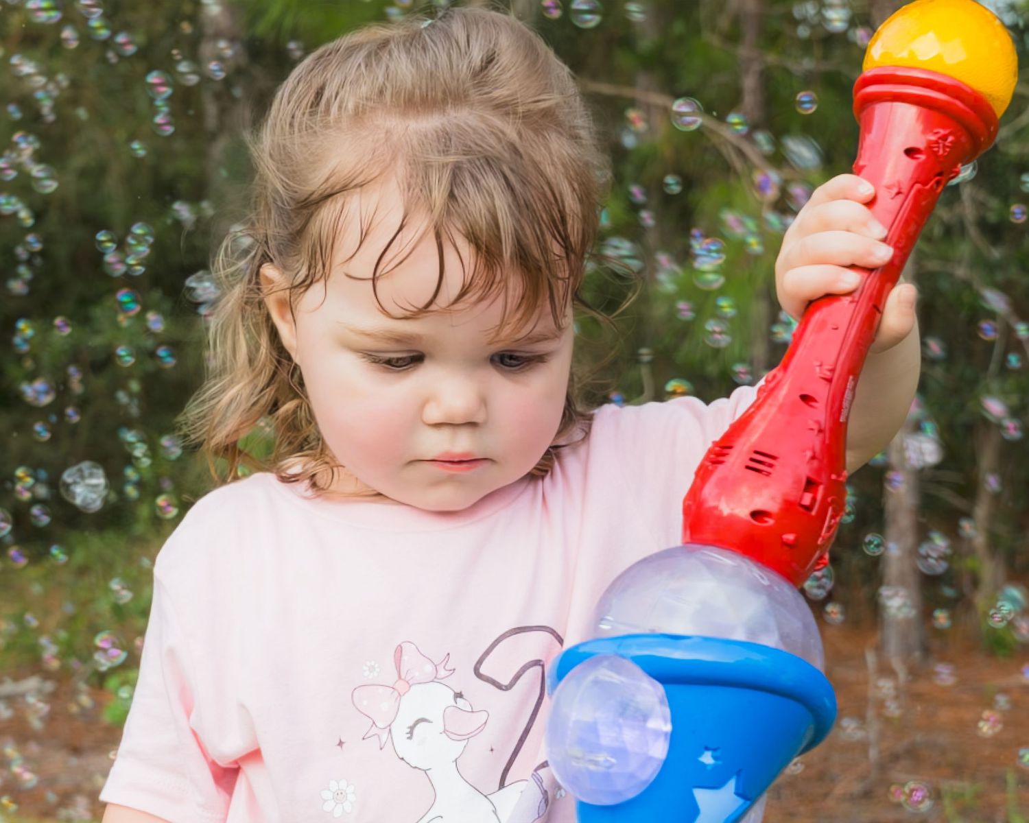 Toddler girl in pink shirt holds colorful bubble wand toy outdoors with bubbles floating around her.