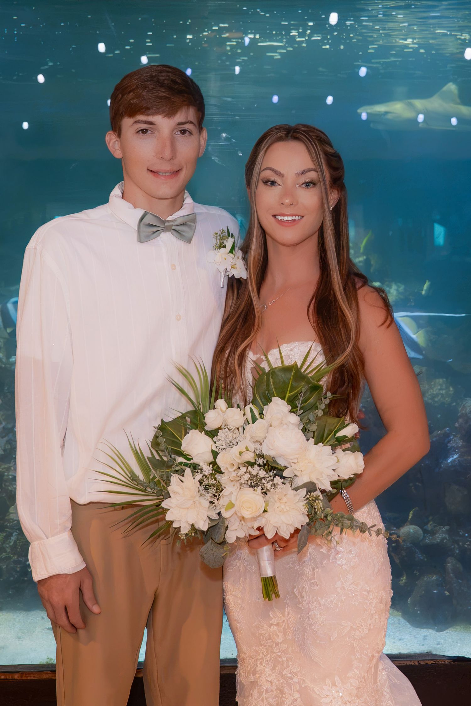 Bride and groom posing in front of large aquarium tank with sharks, bride holding white floral bouquet.