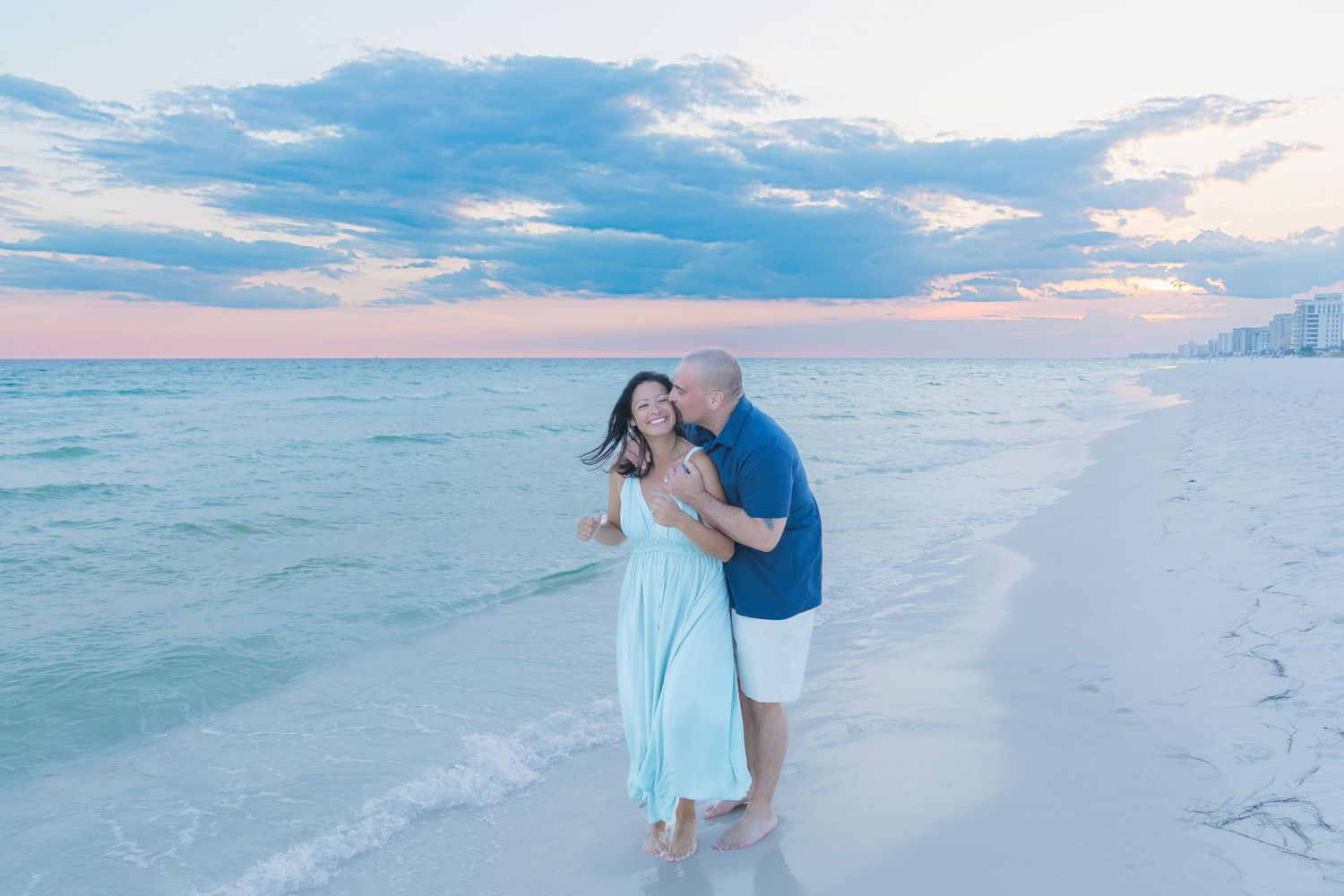 Couple sharing a romantic kiss on a white sand beach at sunset with pastel pink and blue skies.