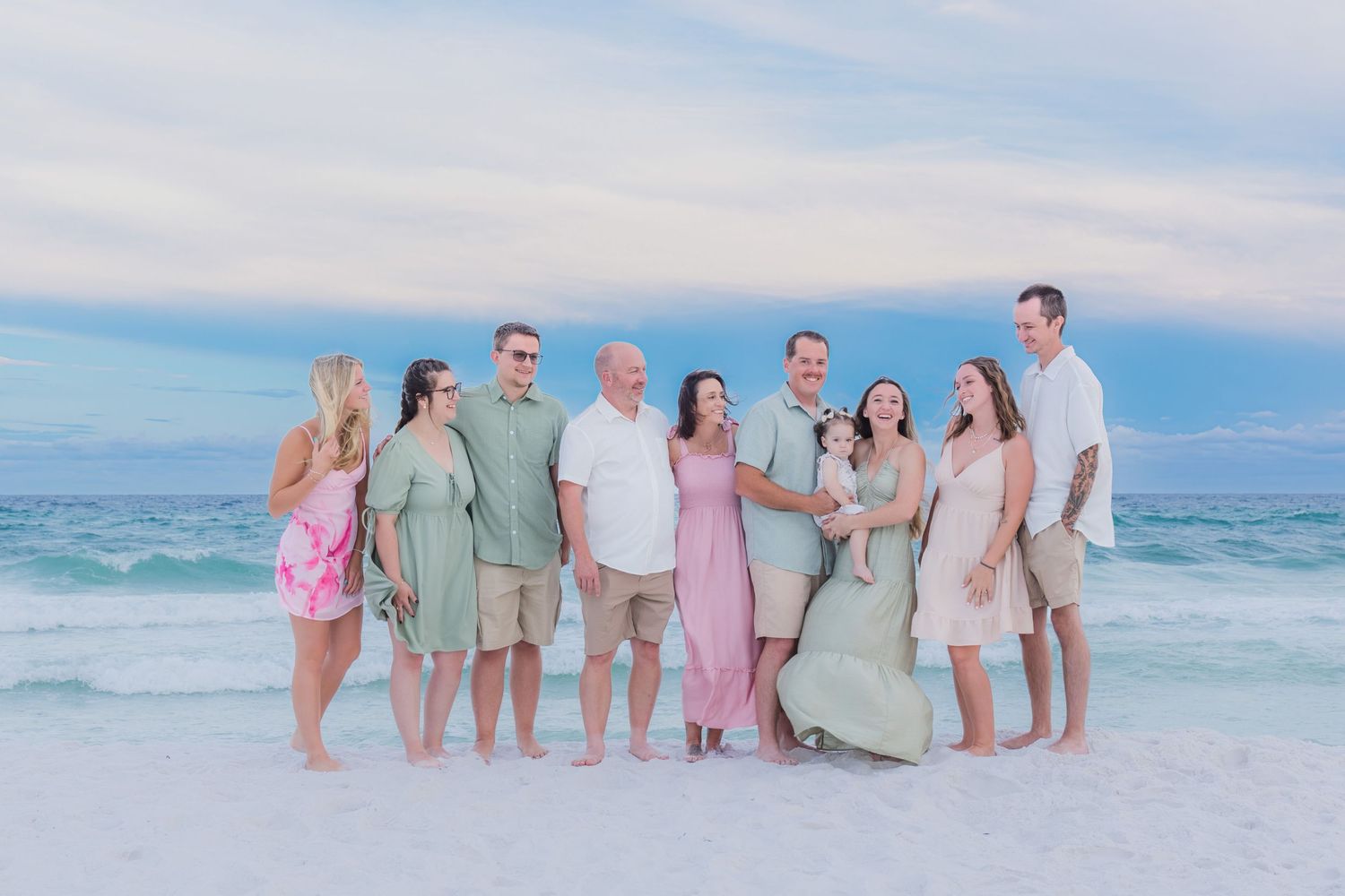 Family group portrait on a sandy beach at sunset, wearing coordinated pastel outfits with ocean waves in the background.