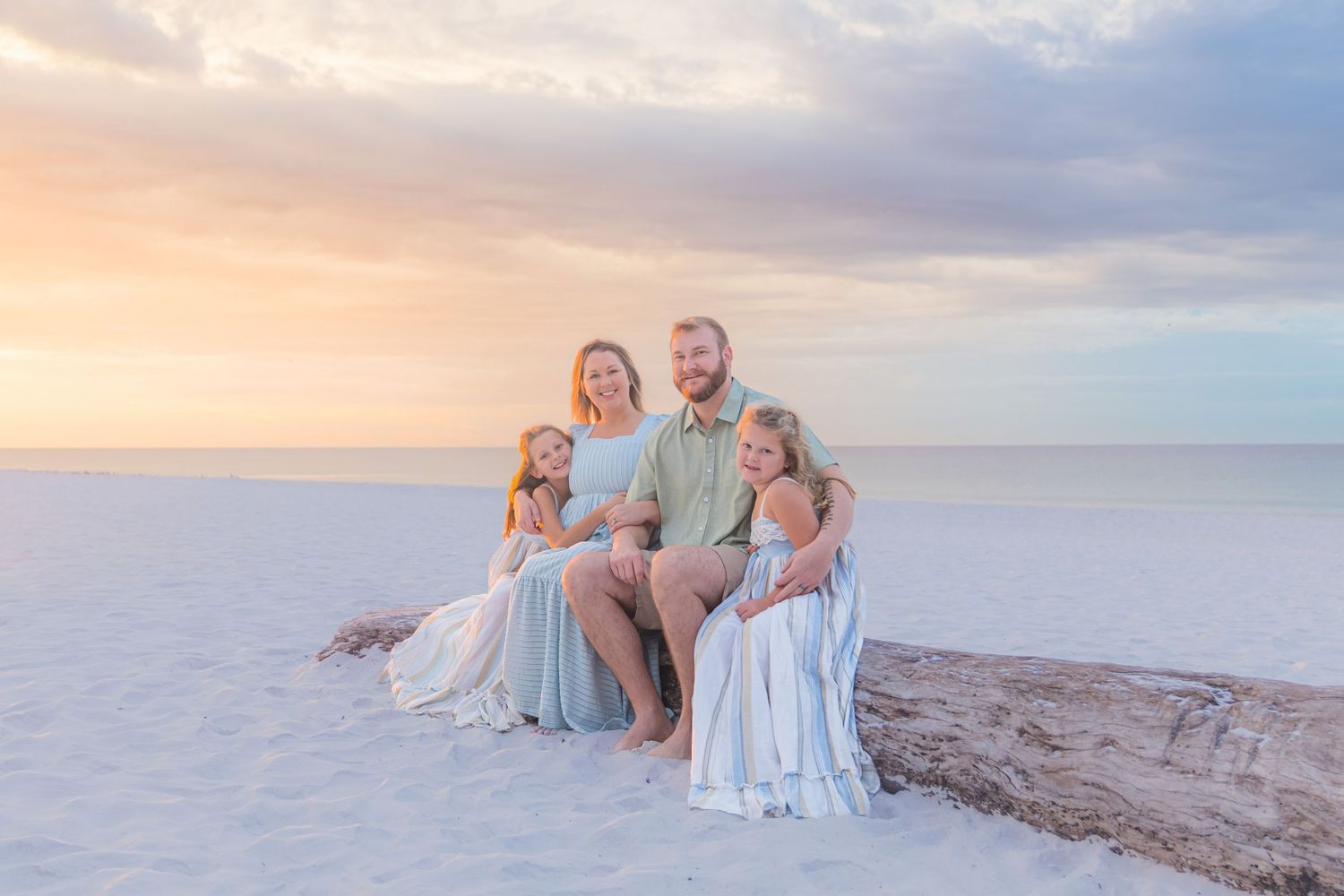 Happy family of four posing on driftwood at sunset on a white sand beach with pastel sky.