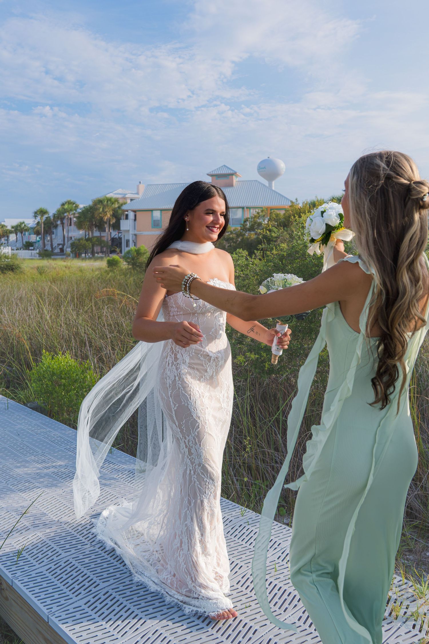 Bride in white lace gown and veil laughs with bridesmaid in mint dress on beach boardwalk.