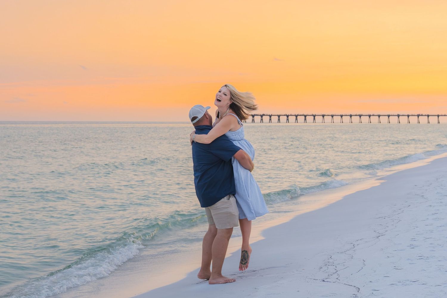 Couple embracing and dancing on a white sand beach at sunset with a pier in the background.