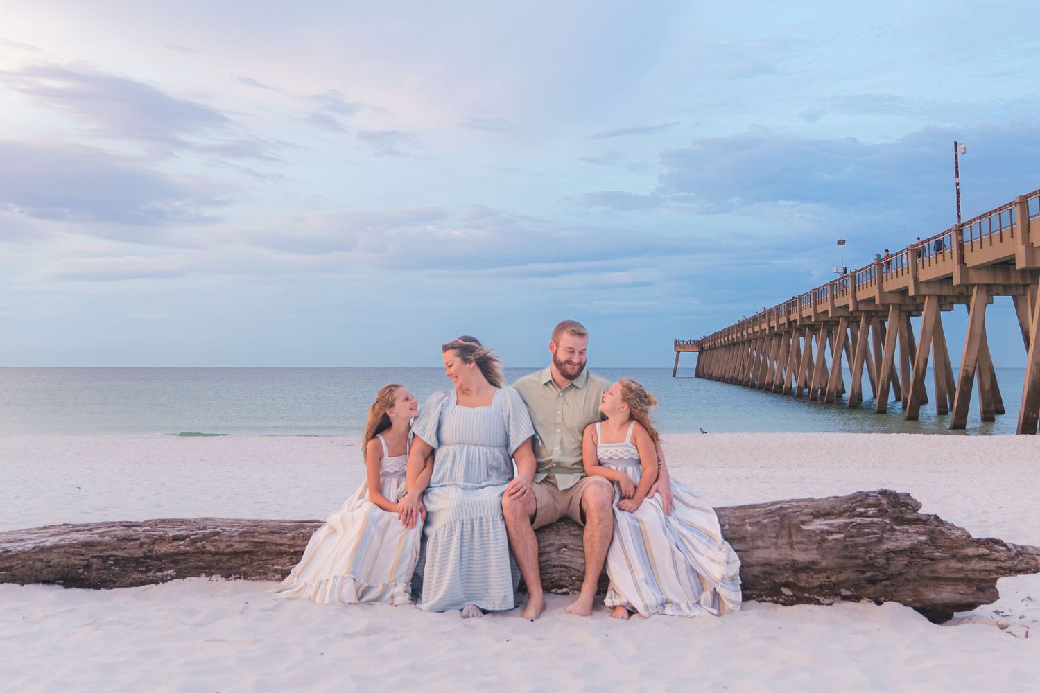 Family of four poses on driftwood at a sandy beach near a pier at sunset in soft blue tones.