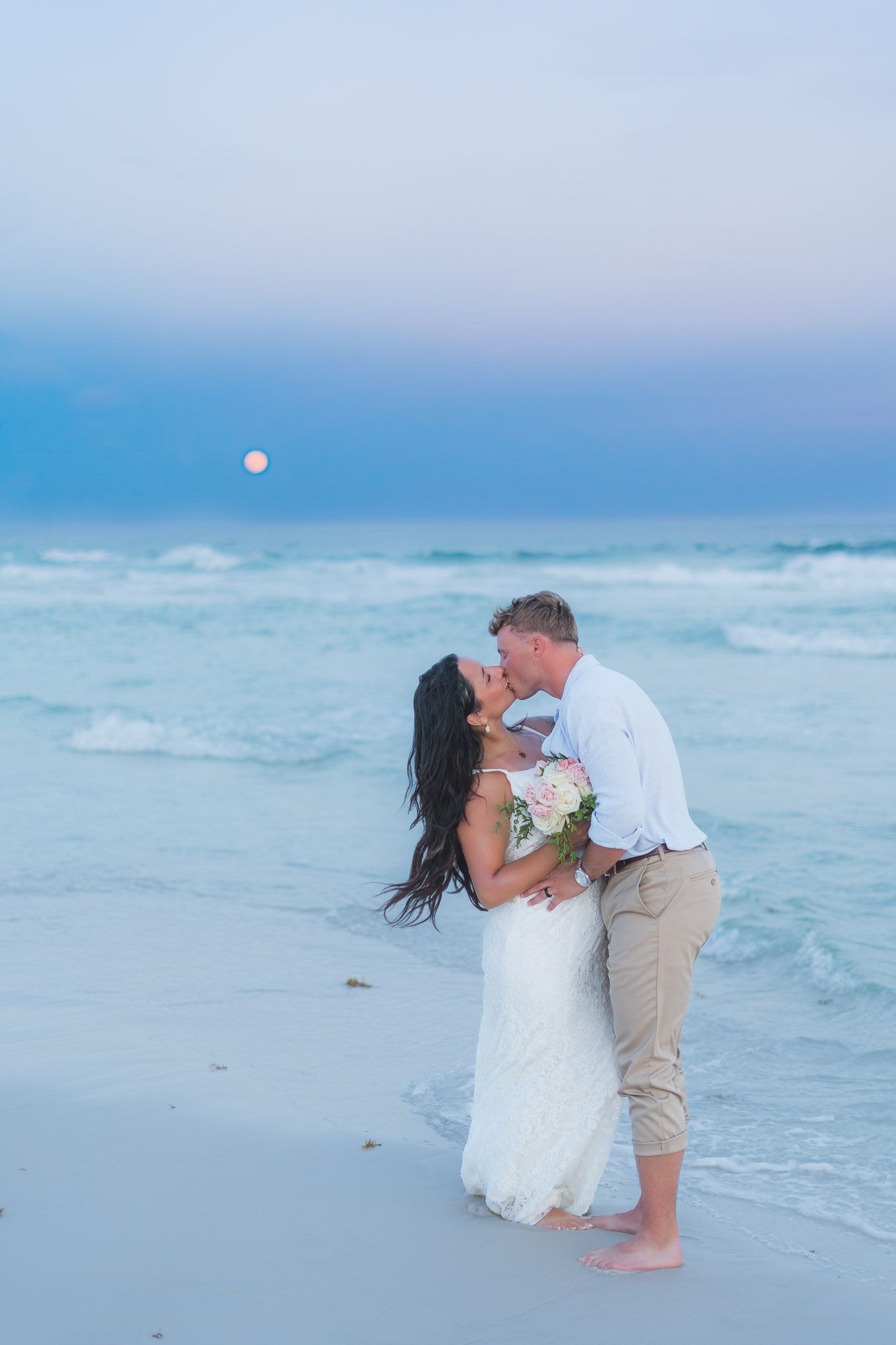 Newlywed couple sharing a romantic beach kiss at sunset, bride in white gown holding bouquet with ocean waves behind them.