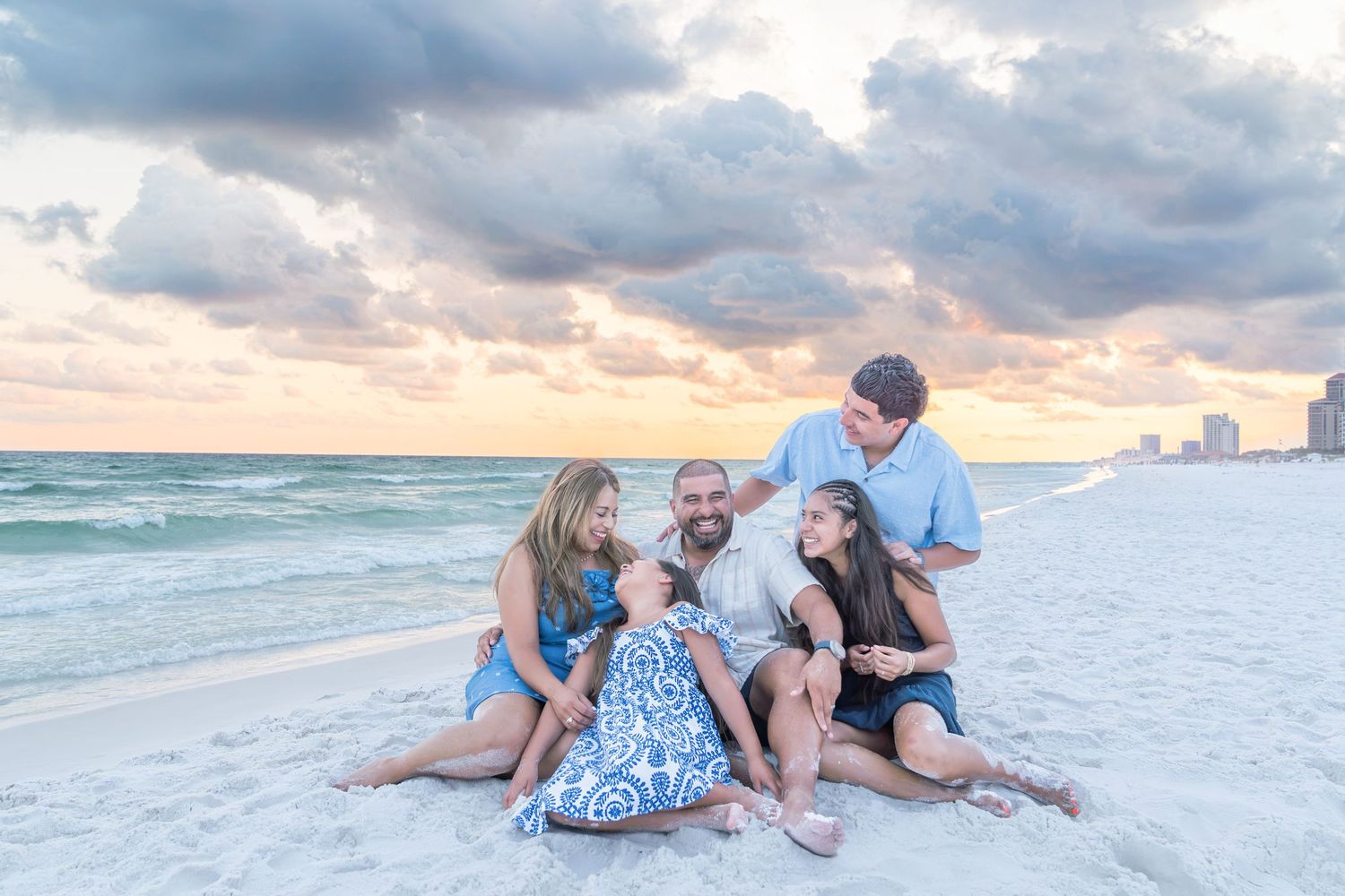 Family of five smiling and sitting together on a white sandy beach at sunset with cloudy skies and ocean in background.
