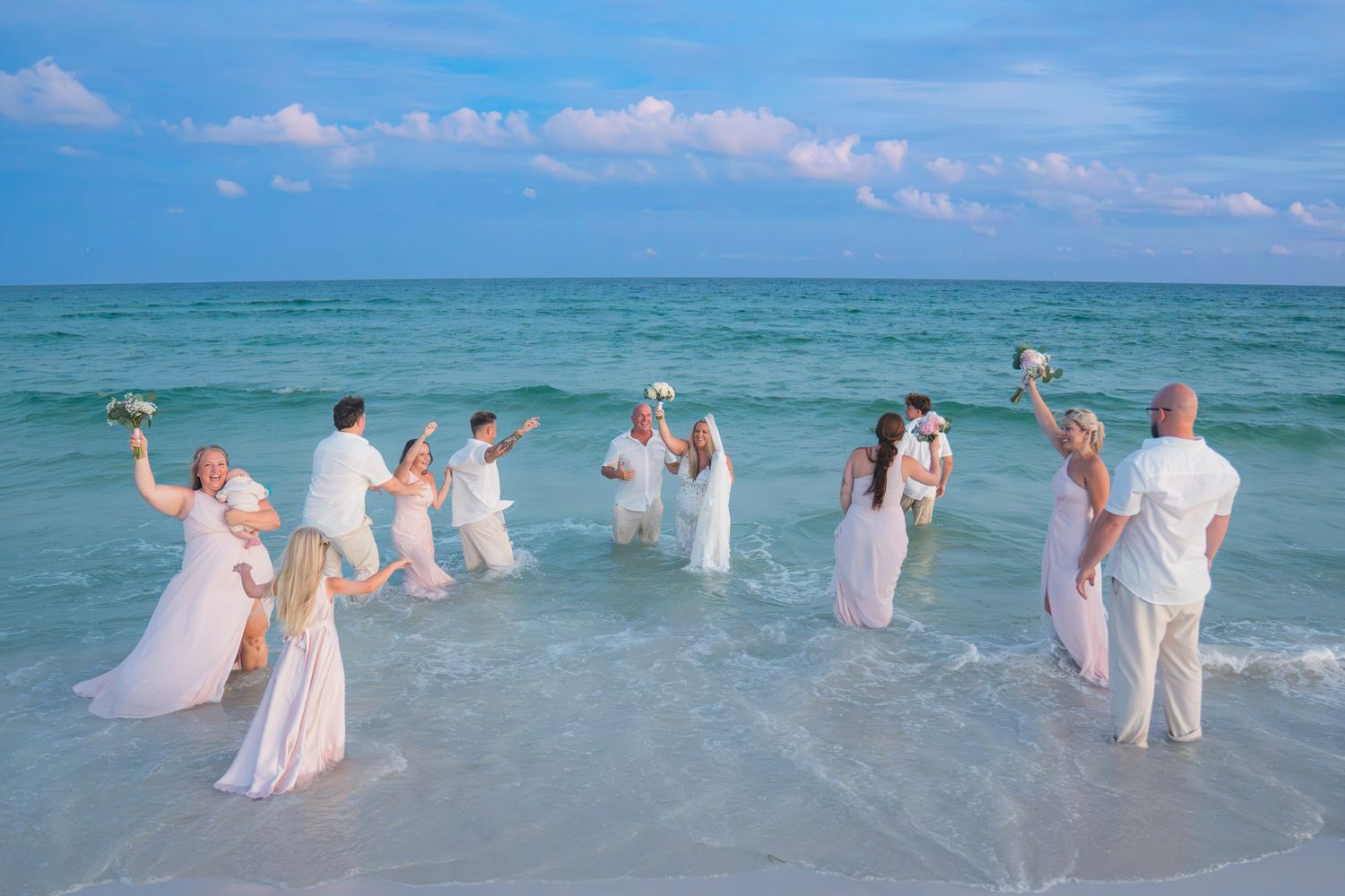 Wedding party celebrates in the ocean waves, dressed in white, holding bouquets on a tropical beach.
