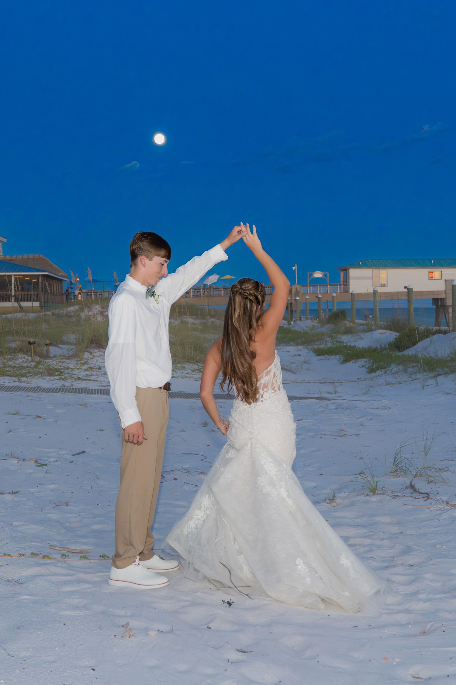 Bride and groom dancing on white sand beach under full moon at twilight in wedding attire.