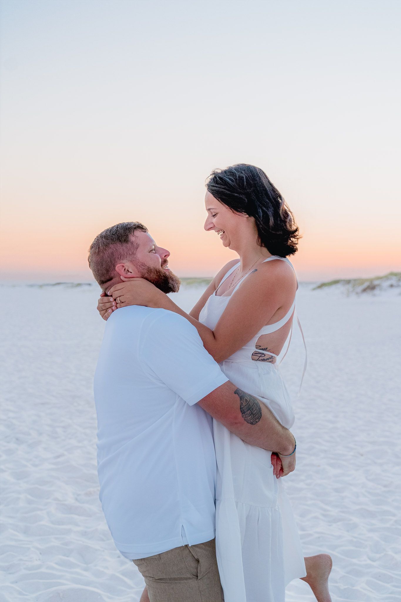 A couple embraces and smiles at each other on a white sand beach during a romantic pastel sunset.