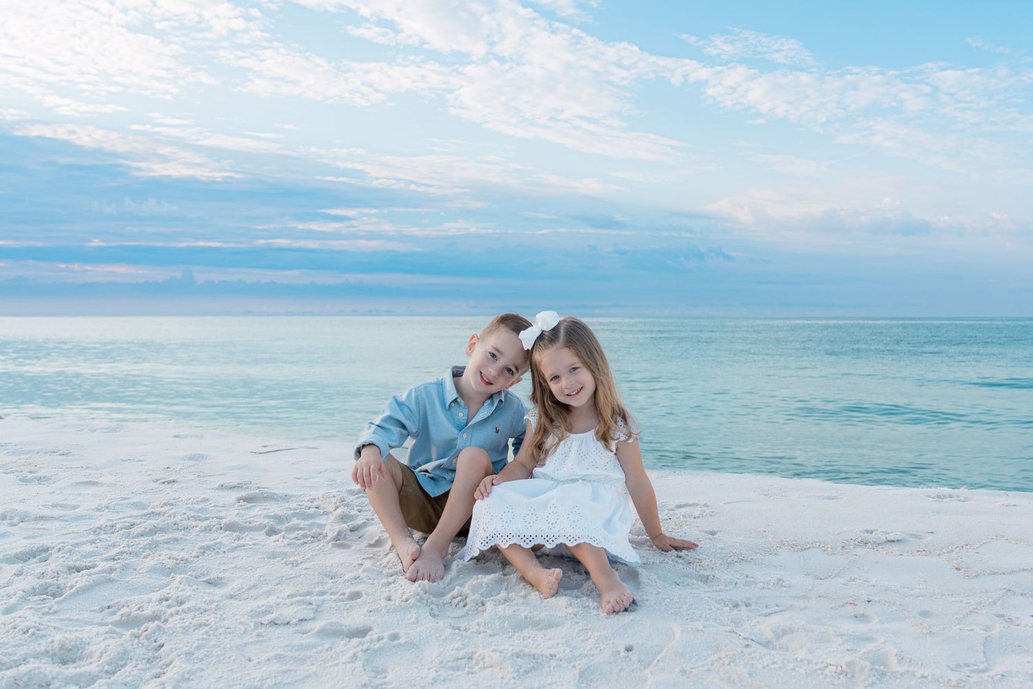 Two children sitting together on a white sandy beach with calm turquoise ocean waters and a pastel cloudy sky.