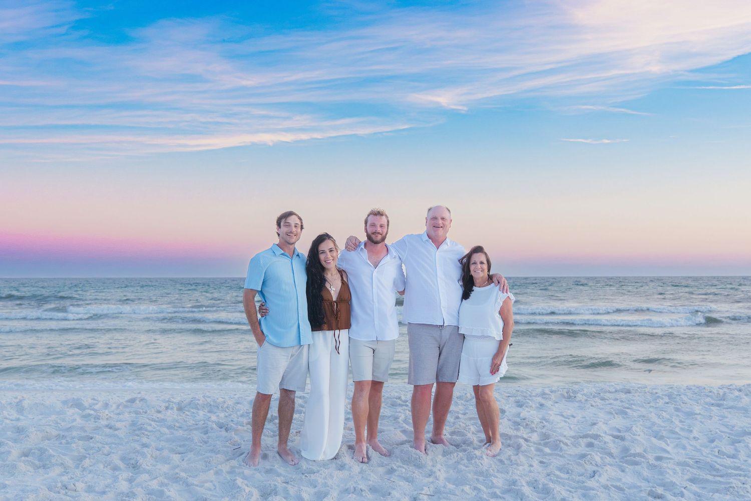 Family of five poses on a white sand beach at sunset with pastel skies and ocean waves in the background.