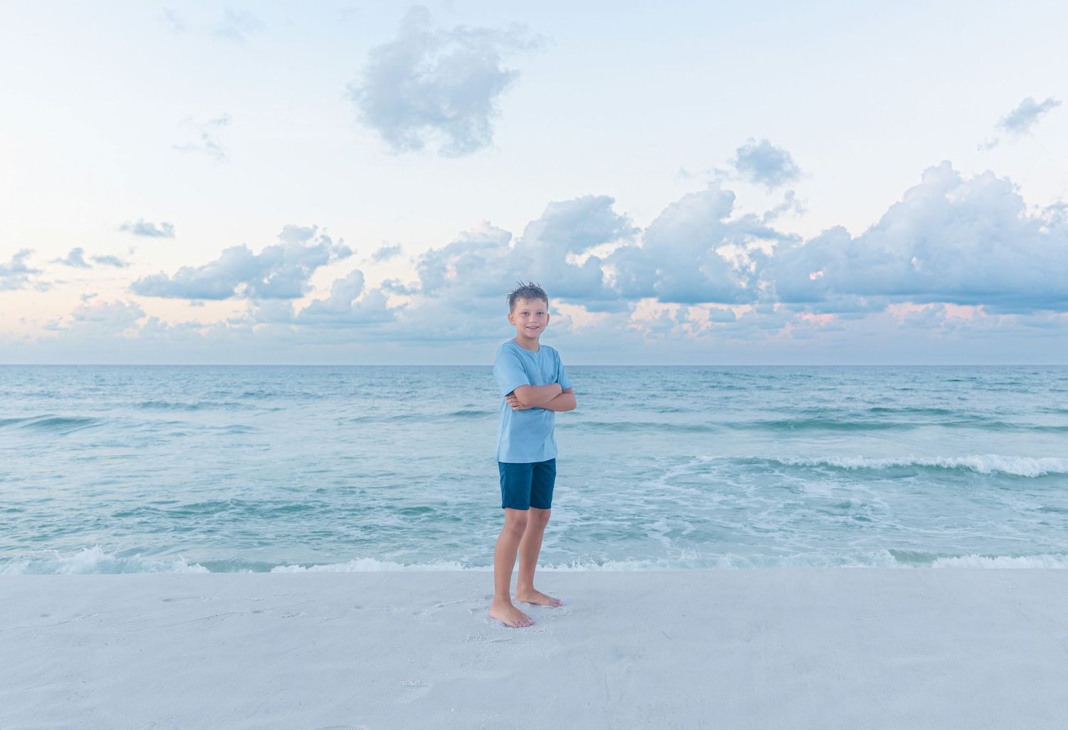 A young boy stands with arms crossed on a white sand beach, smiling at sunset with calm ocean waves behind him.