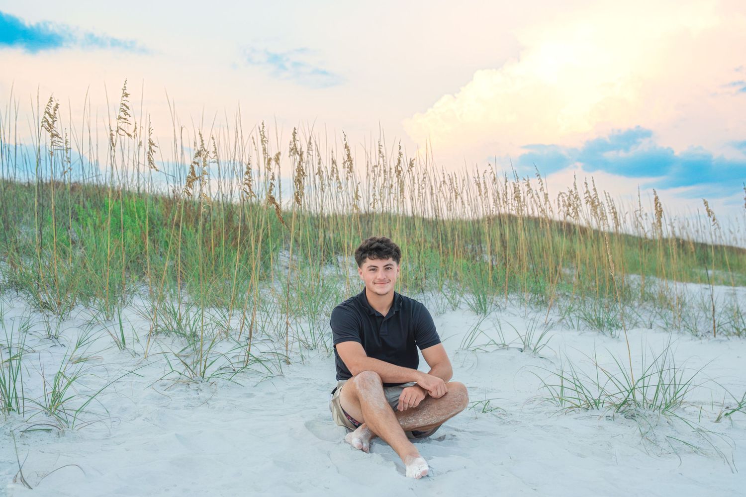 Young man sitting cross-legged on white sand dunes with sea oats and pastel sunset sky at the beach.